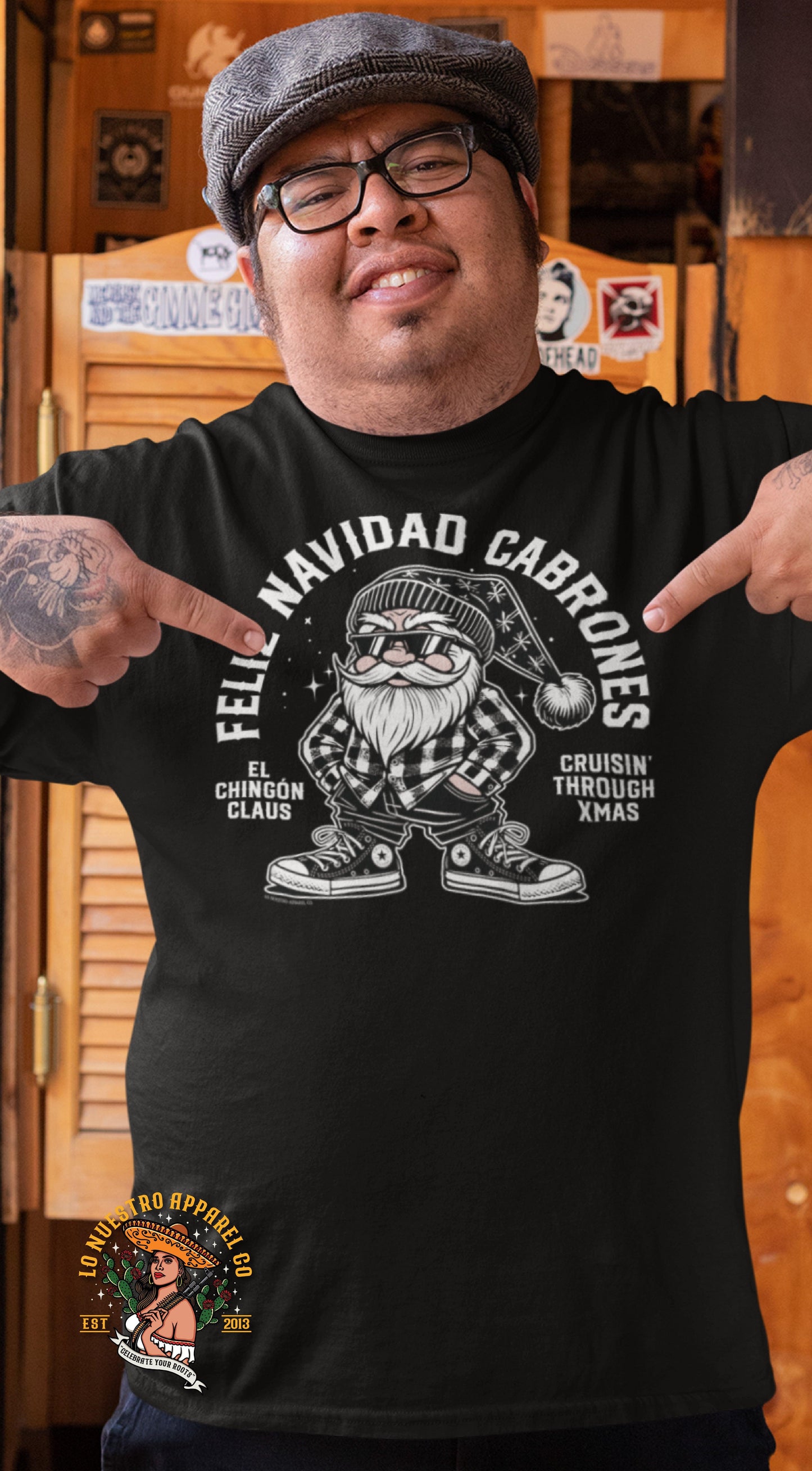 Man in a rustic bar wearing a black 'Feliz Navidad Cabrones' t-shirt with El Chingón Claus and 'Cruisin’ through Xmas.' The man is pointing at the design proudly.