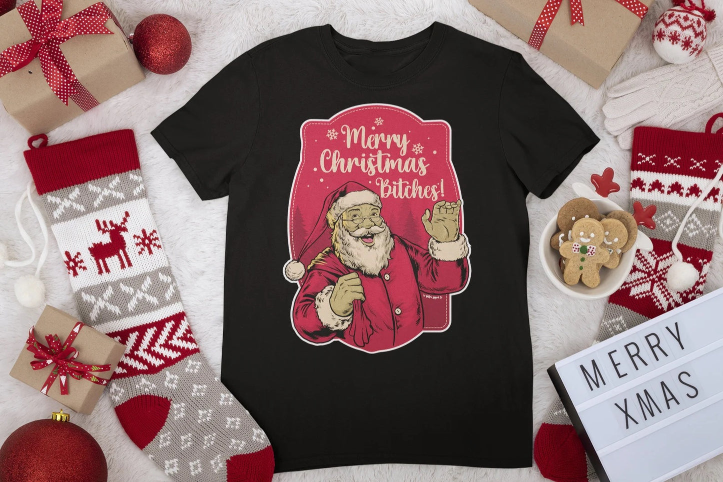 Flat lay of a "Merry Christmas Bitches" unisex shirt surrounded by holiday decorations, showcasing the playful Santa design.