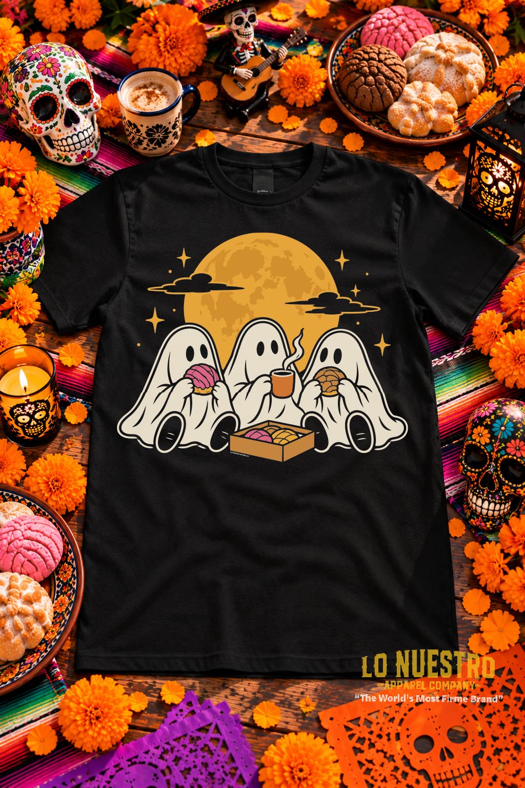 Black crewneck t-shirt laid flat on a rustic wooden table surrounded by colorful sugar skulls, orange marigolds, papel picado, Mexican pan dulce, coffee, and Día de los Muertos decorations.