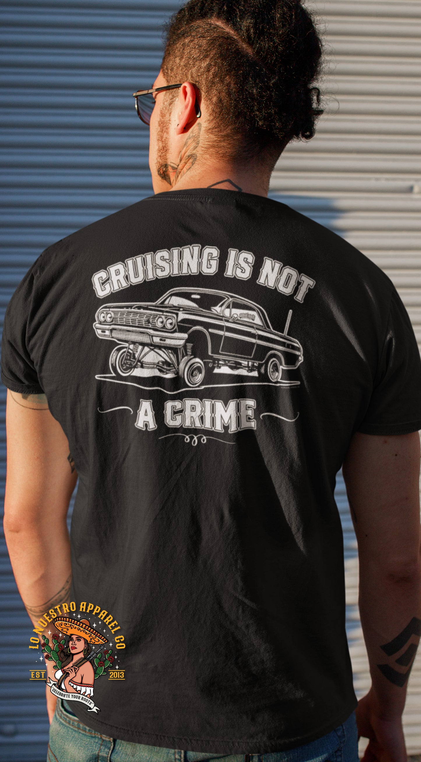 A person wearing a black T-shirt with the back design featuring a bold print of a classic lowrider car and the phrase "Cruising Is Not a Crime.