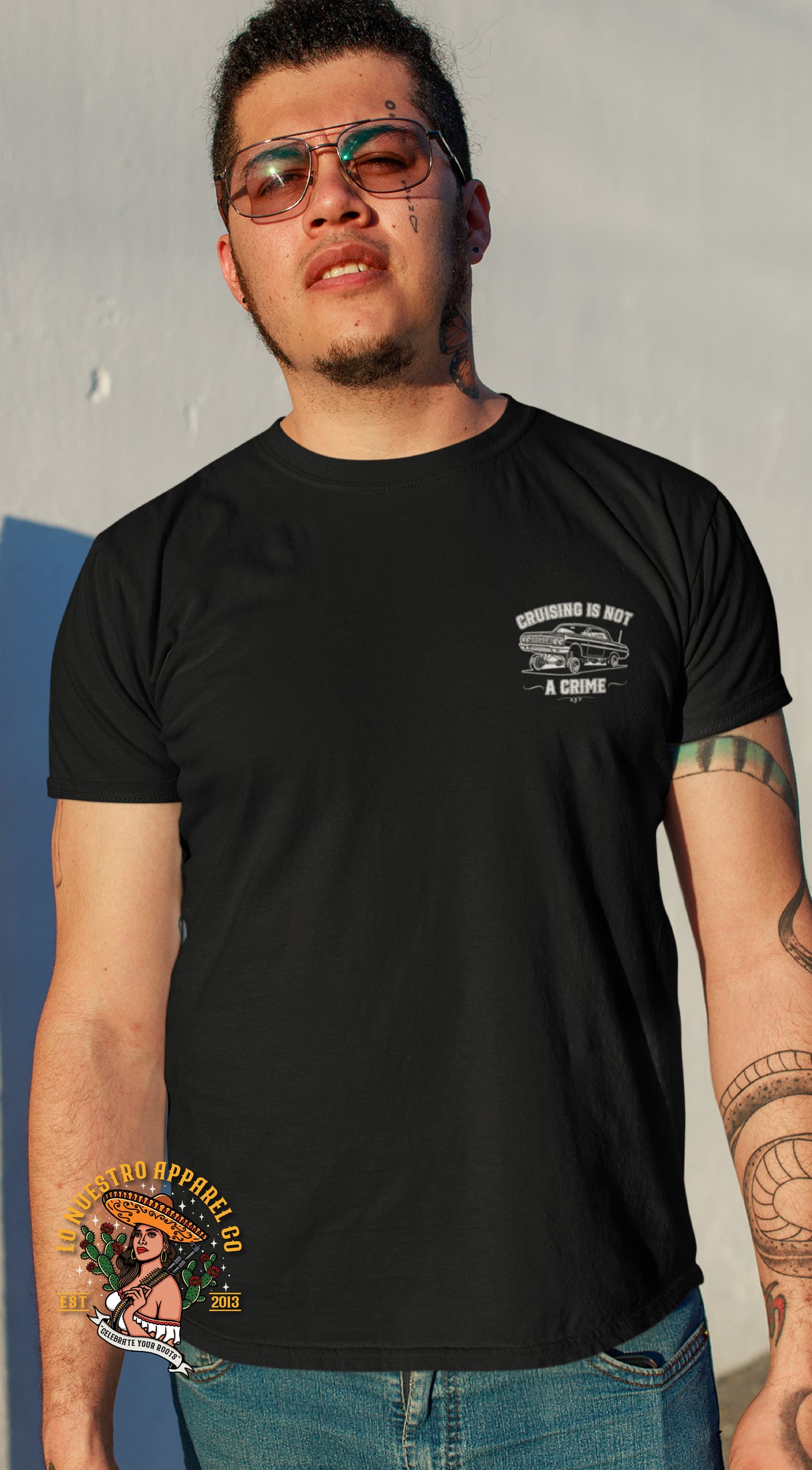 A man with tattoos is wearing the black T-shirt featuring a small front chest graphic of a lowrider car with the phrase "Cruising Is Not a Crime.