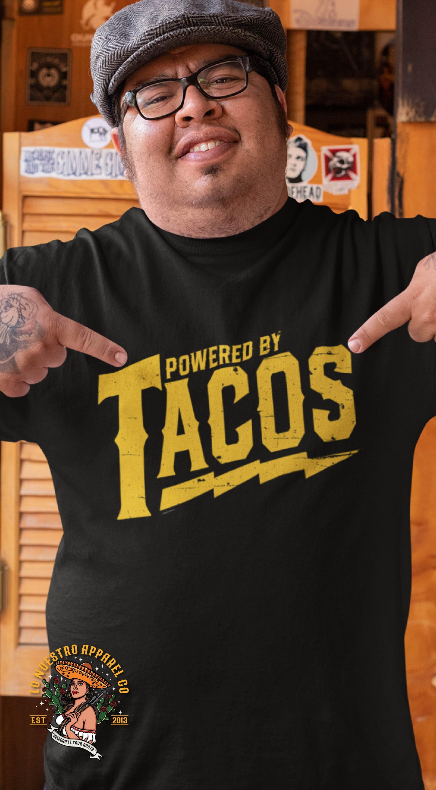Man pointing at his black graphic t-shirt with bold yellow 'Powered by Tacos' text, showing taco pride.