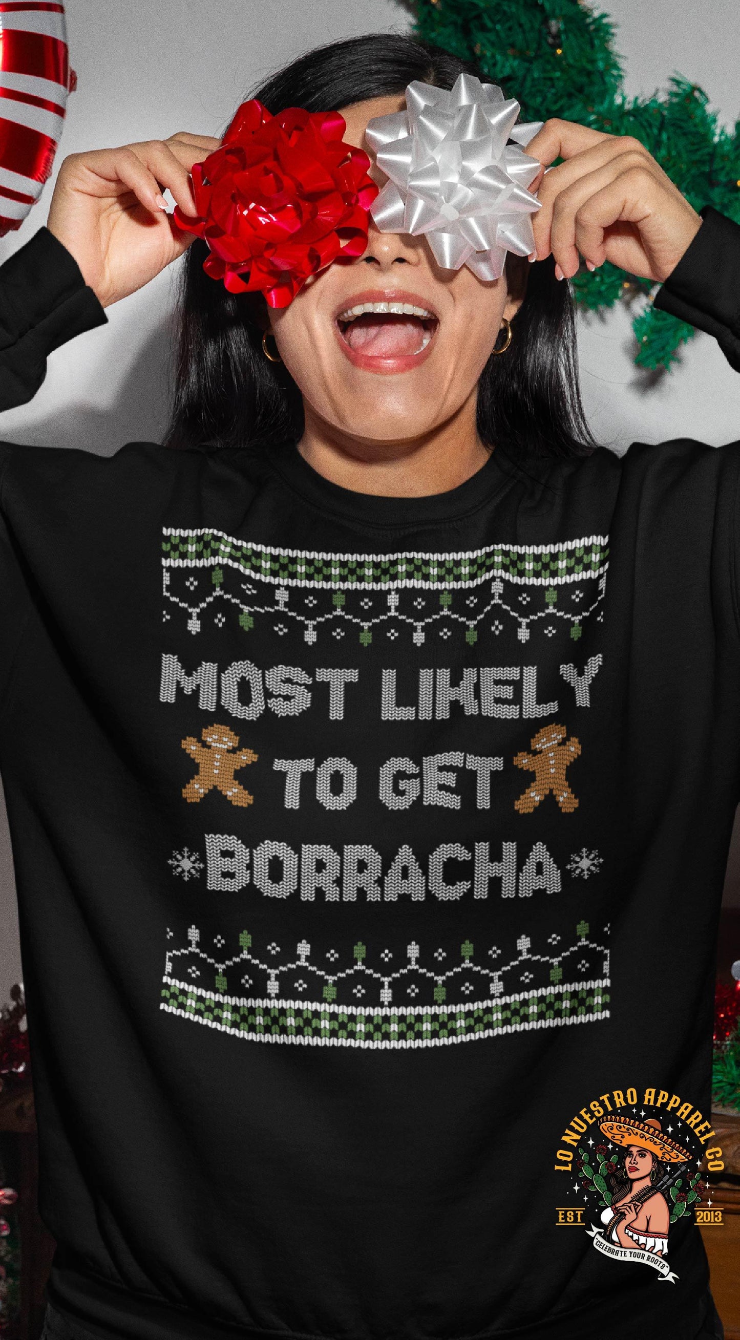Woman celebrating Christmas with ribbons over her eyes, wearing a black sweater that says 'Most Likely to Get Borracha' with gingerbread men design