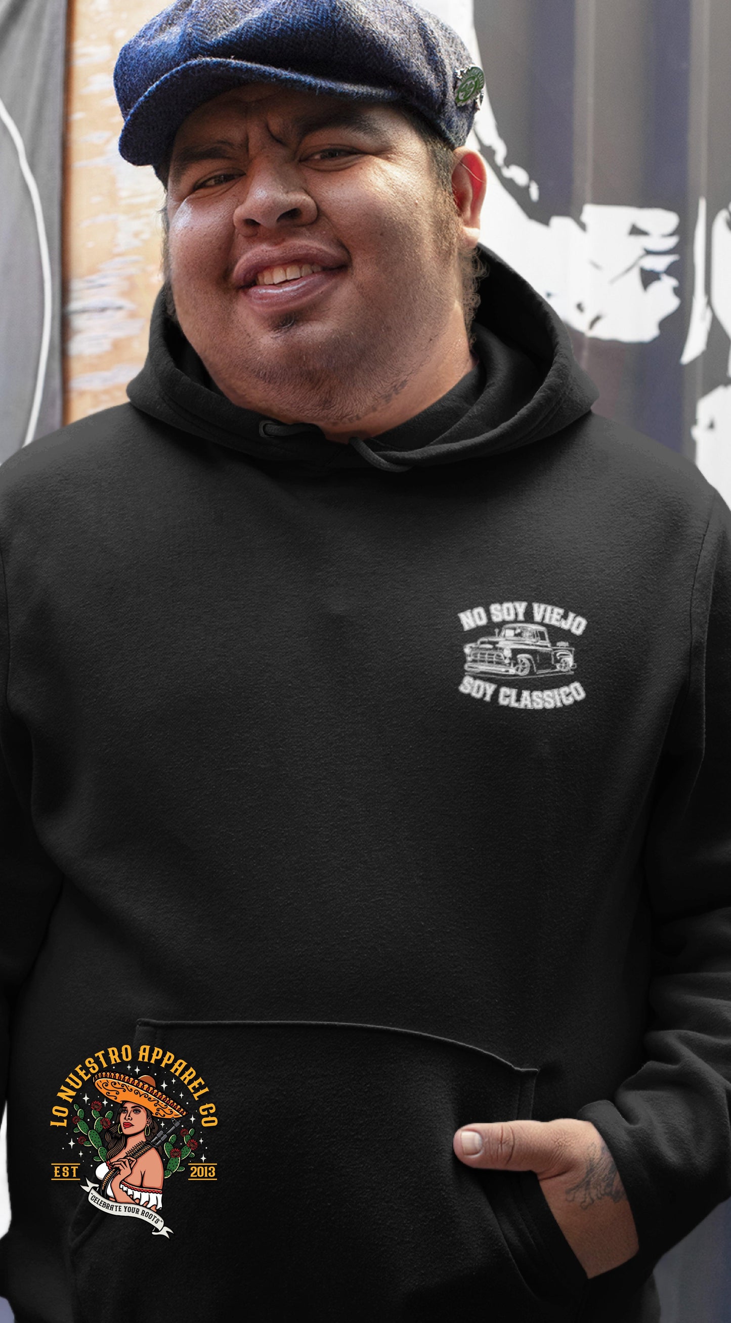 Smiling man wearing a black hoodie with a “No Soy Viejo, Soy Classico” graphic over the chest, standing outdoors in front of a mural.