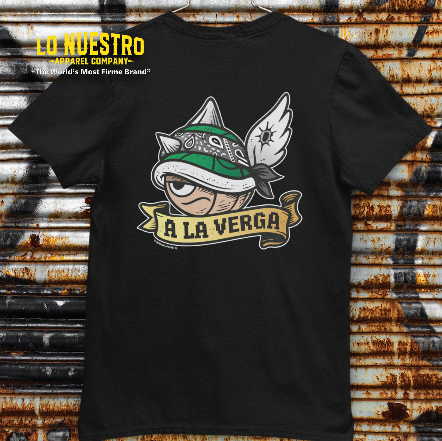 Back view of A La Verga t-shirt hanging on a rusty metal wall, showcasing a green shell graphic with wings and a banner; funny Latino shirt for gamers.