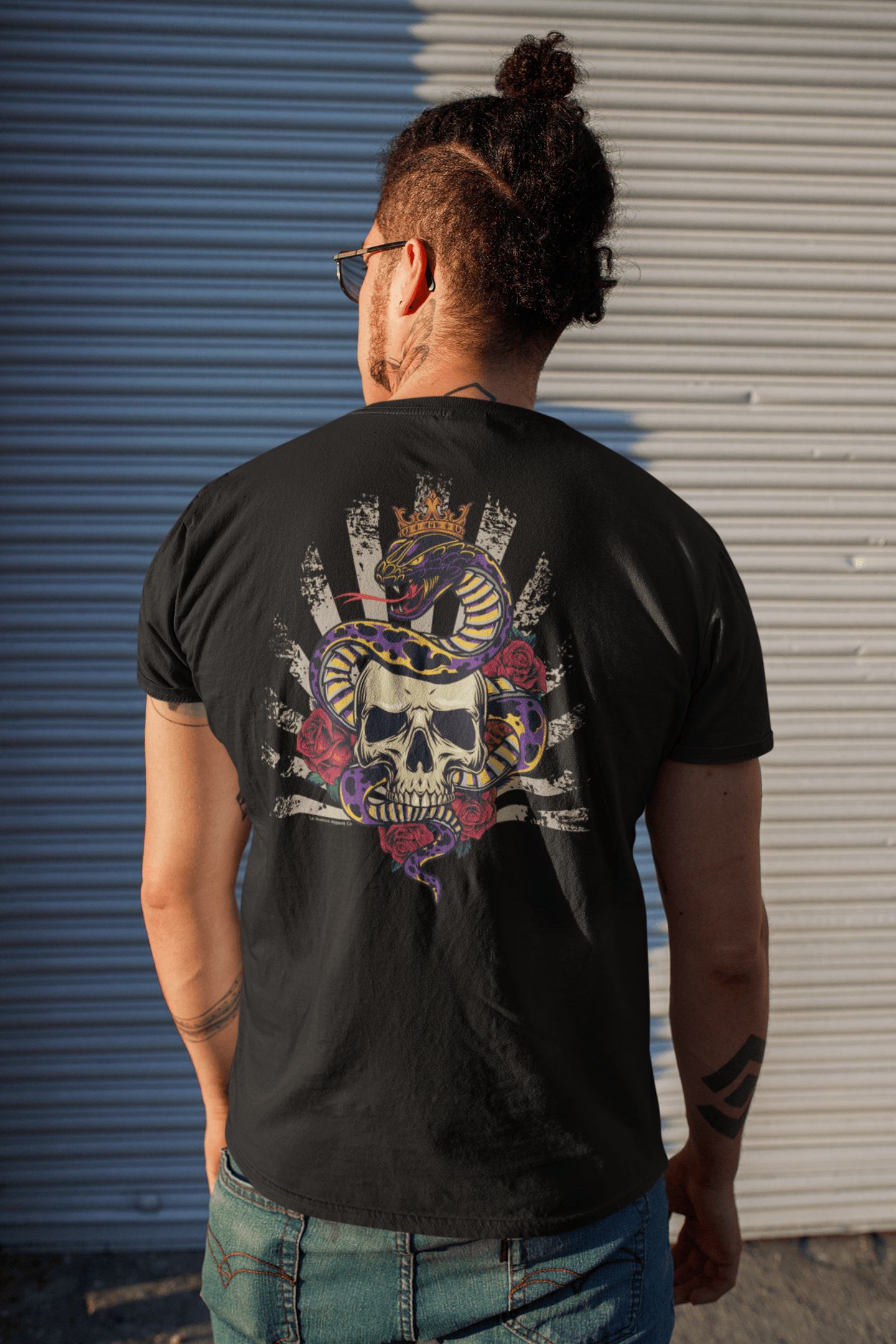 Man wearing the G.O.A.T. T-Shirt, showing the back graphic of a crowned snake wrapped around a skull, standing on the street with a bun hairstyle.