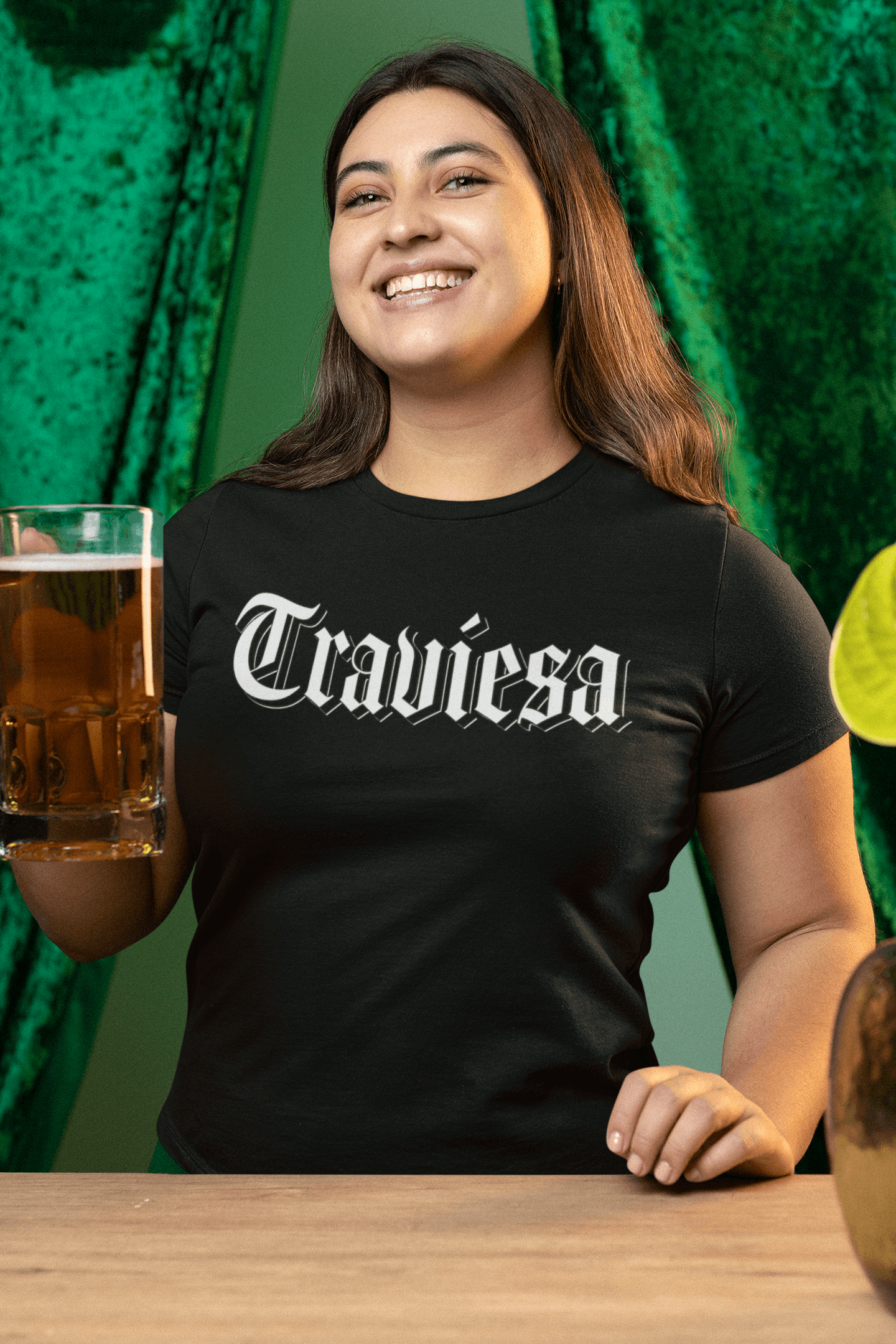 A smiling woman wearing a black relaxed t-shirt with 'Traviesa' in Gothic-style lettering, holding a beer.