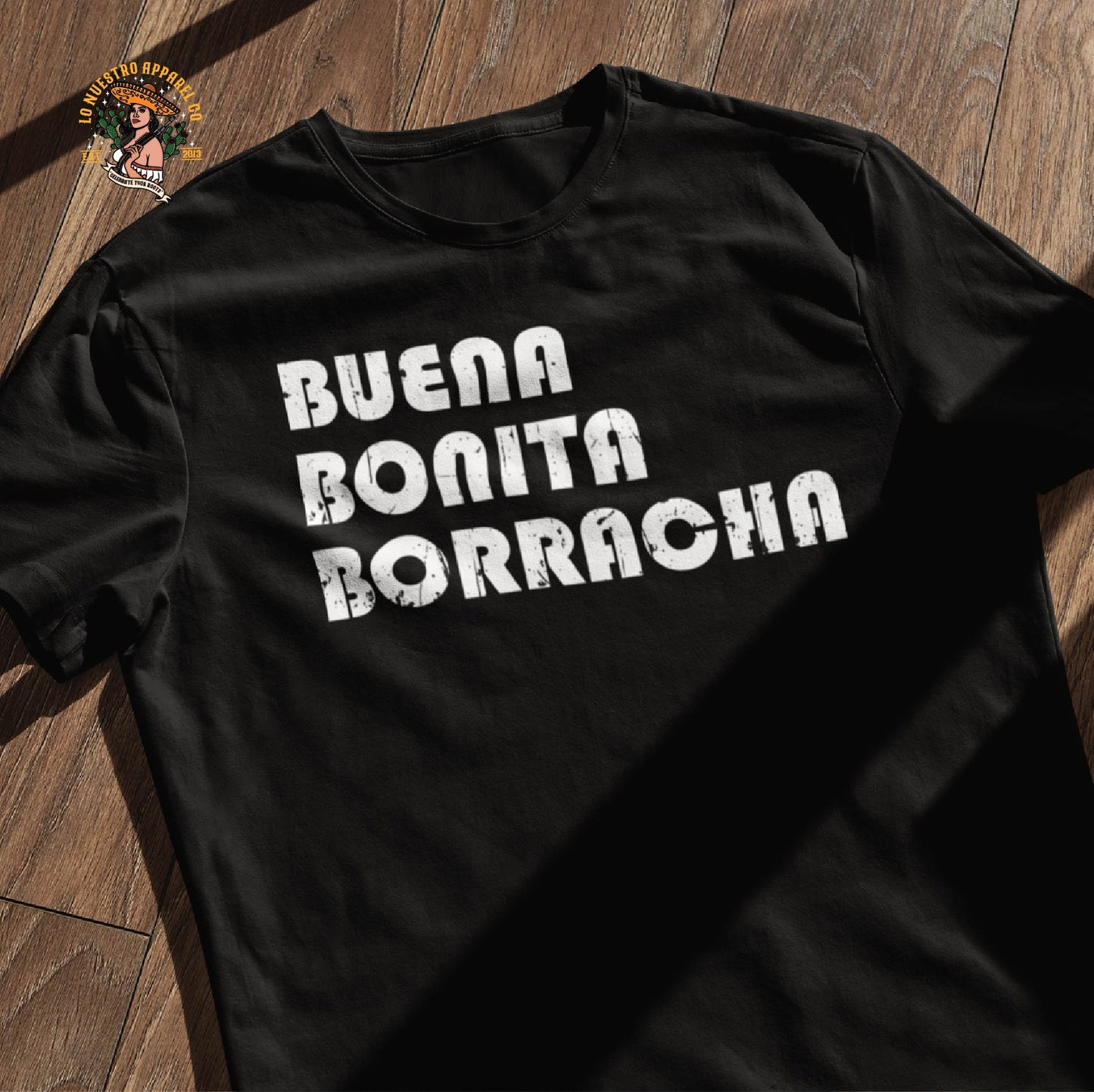 A black tshirt with the Buena Bonita Borracha distressed text on a wooden floor. Part of our funny t shirts for women line.