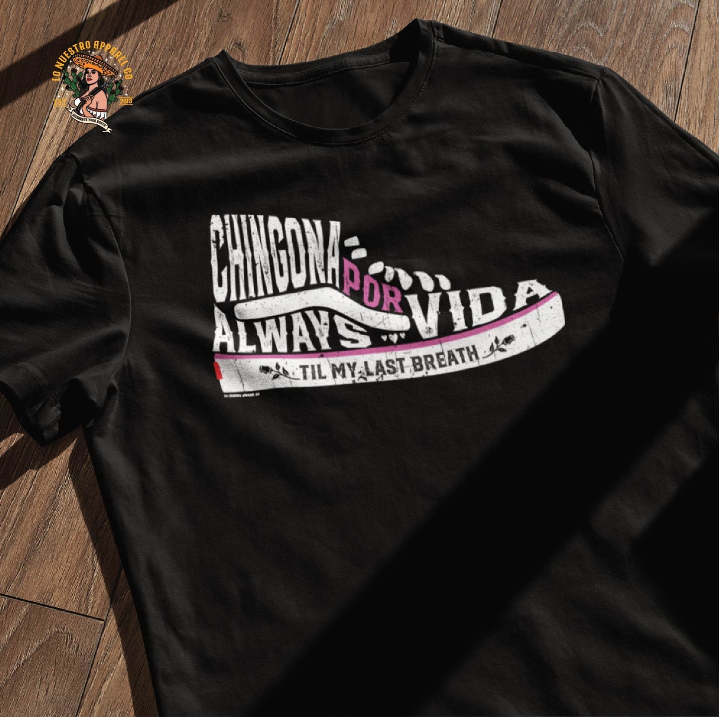 Flat lay of the Chingona Por Vida Chicana t-shirt on a wooden floor. Bold and empowering design for Chicana shirts collection.
