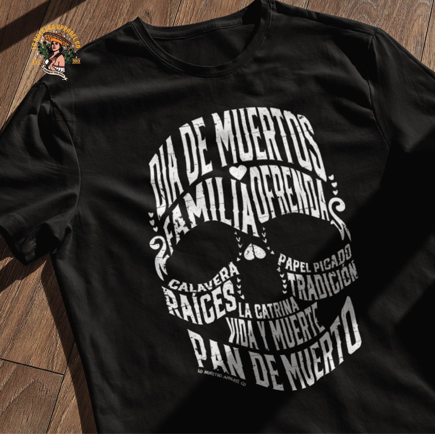 Black Dia de los Muertos calavera shirt laid on a wooden surface, featuring a detailed calligram skull design inspired by Chicano shirts and Day of the Dead culture.
