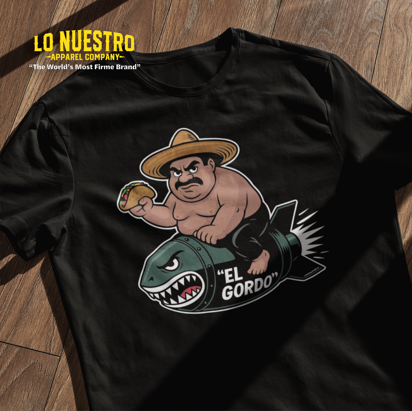 A black t-shirt with a graphic of a chubby man riding a shark-faced bomb labeled El Gordo, holding a taco and wearing a sombrero, displayed on a wooden floor with sunlight casting window shadows.