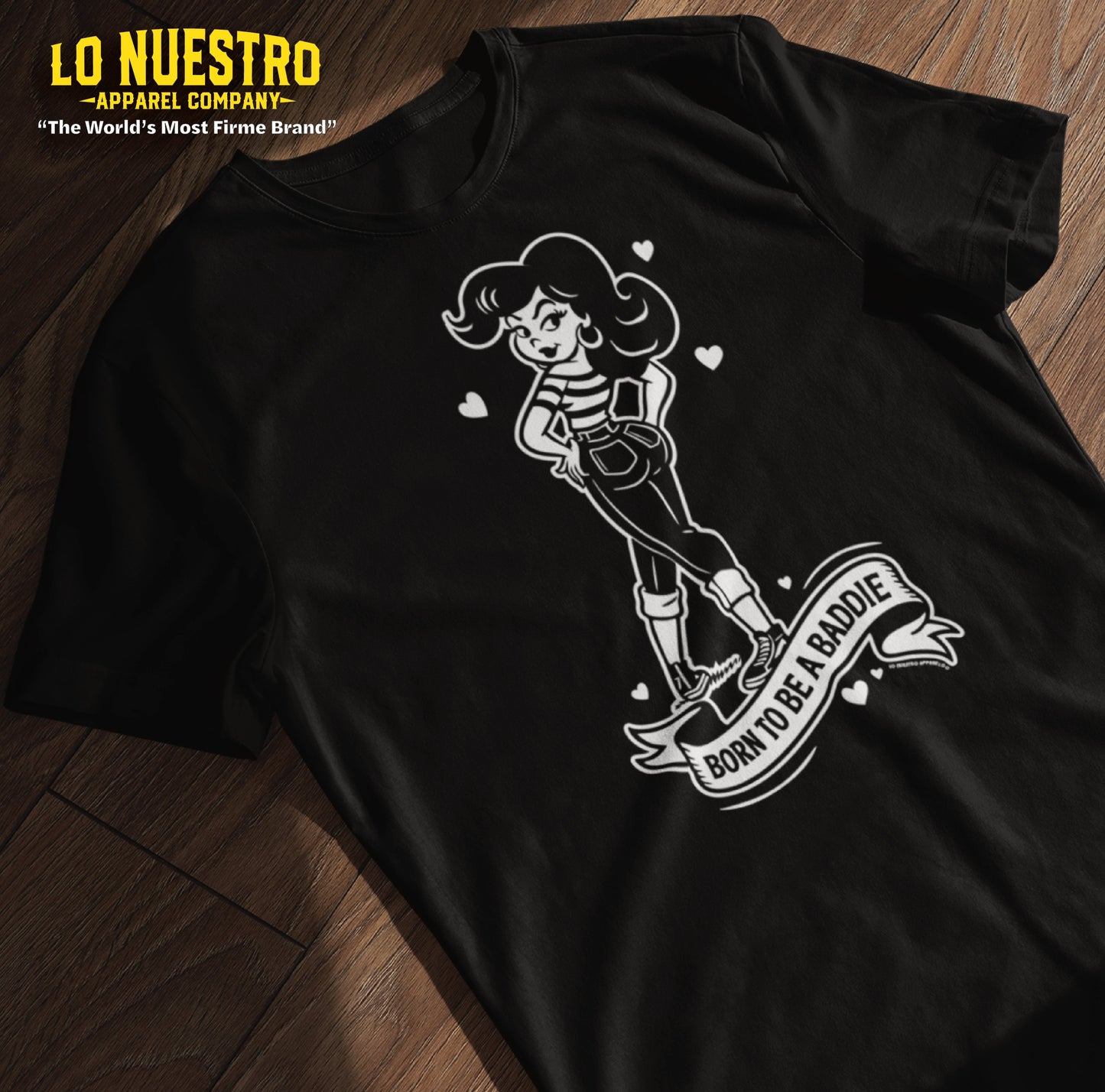 Flatlay of a black Chicana shirt on a wooden floor featuring a vintage cartoon-style girl with the phrase Born to Be a Baddie.
