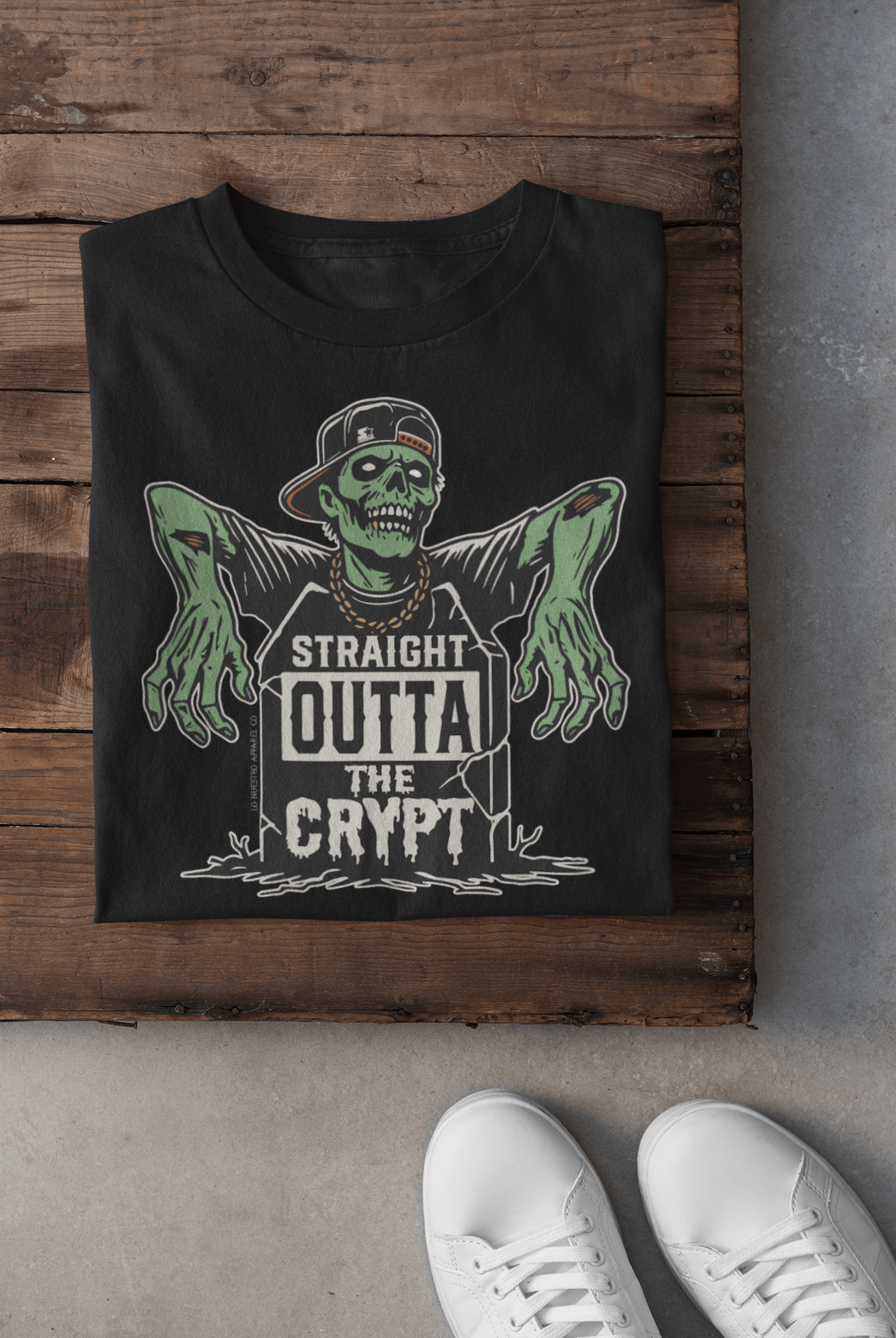 Folded black t-shirt featuring a 'Straight Outta the Crypt' zombie design with desaturated colors on a wooden surface