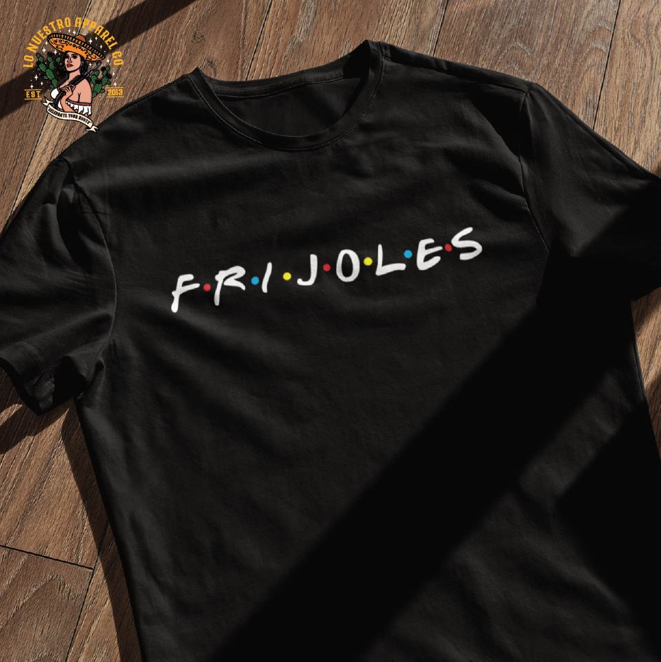 A black Frijoles Tshirt on a wooden floor and Lo Nuestro Apparel Co logo. This is a parody tshirt of the FRIENDS logo with a Chicano Shirt and Chicano Clothing twist. This is a Funny Mexican T Shirt.