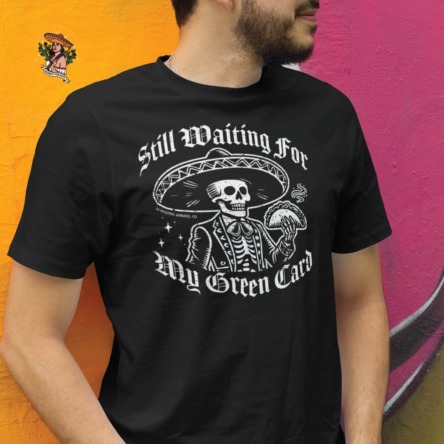 Funny Mexican shirt featuring a skeleton mariachi in a sombrero holding a taco with Old English text that reads Still Waiting for My Green Card. This Chicano clothing design is worn by a male model against a colorful mural background. Perfect for those looking for funny shirts for men.
