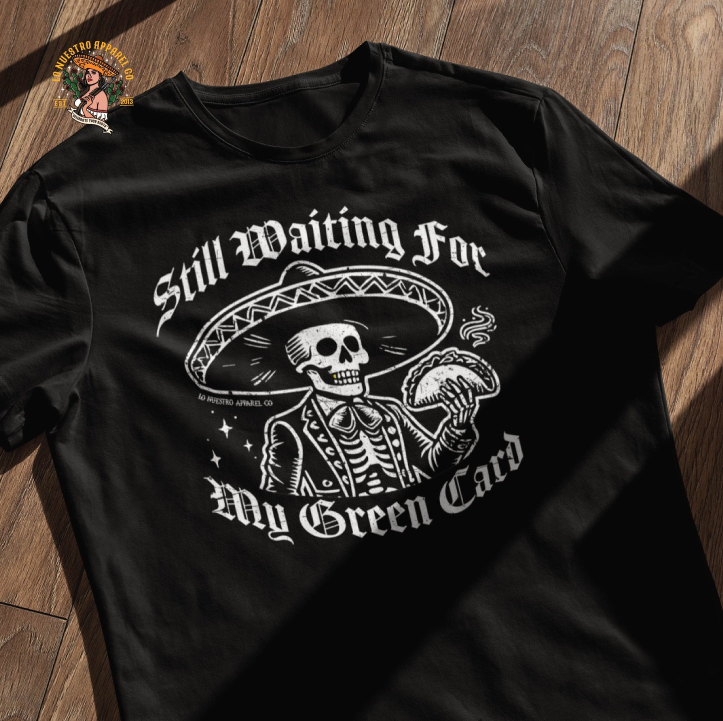 Flat lay of a funny Mexican shirt with a skeleton mariachi design, holding a taco and wearing a sombrero. The Old English text reads Still Waiting for My Green Card. This Chicano clothing piece blends humor and cultura, making it a great addition to any collection of funny shirts for men.