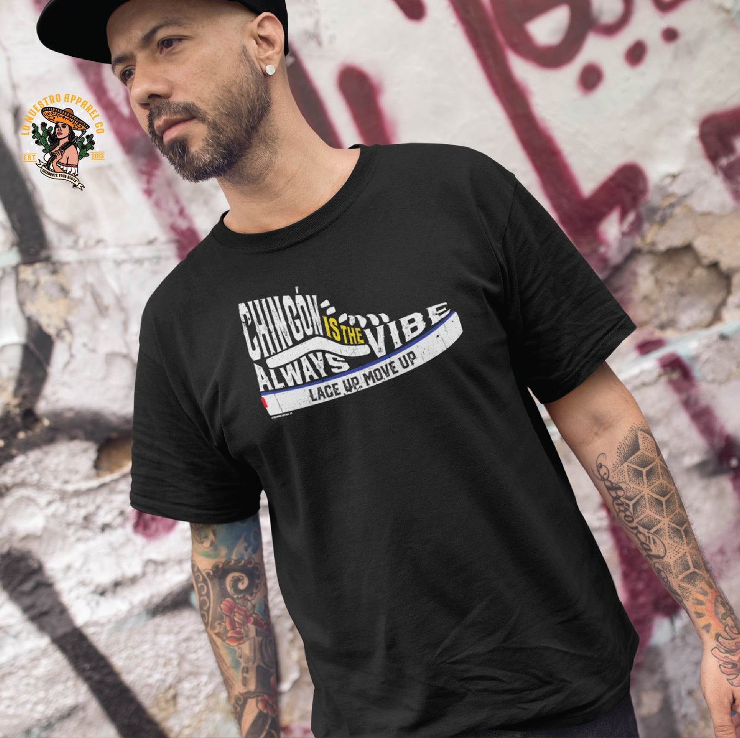 Tattooed man wearing a black Chingón Is The Vibe shirt, a bold Chicano shirt with a sneaker-inspired design, standing in front of urban graffiti.
