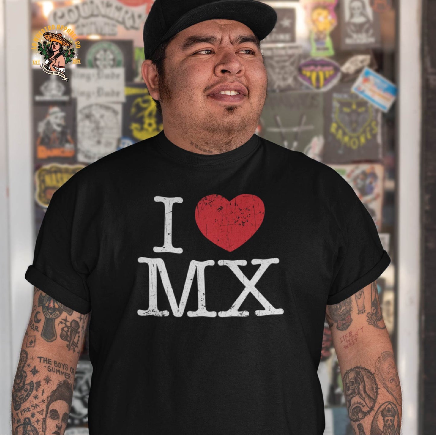 A man with tattooed arms wearing an I Love MX Mexican shirt, standing in front of a sticker-covered glass door. A bold way to rep Mexico with style.