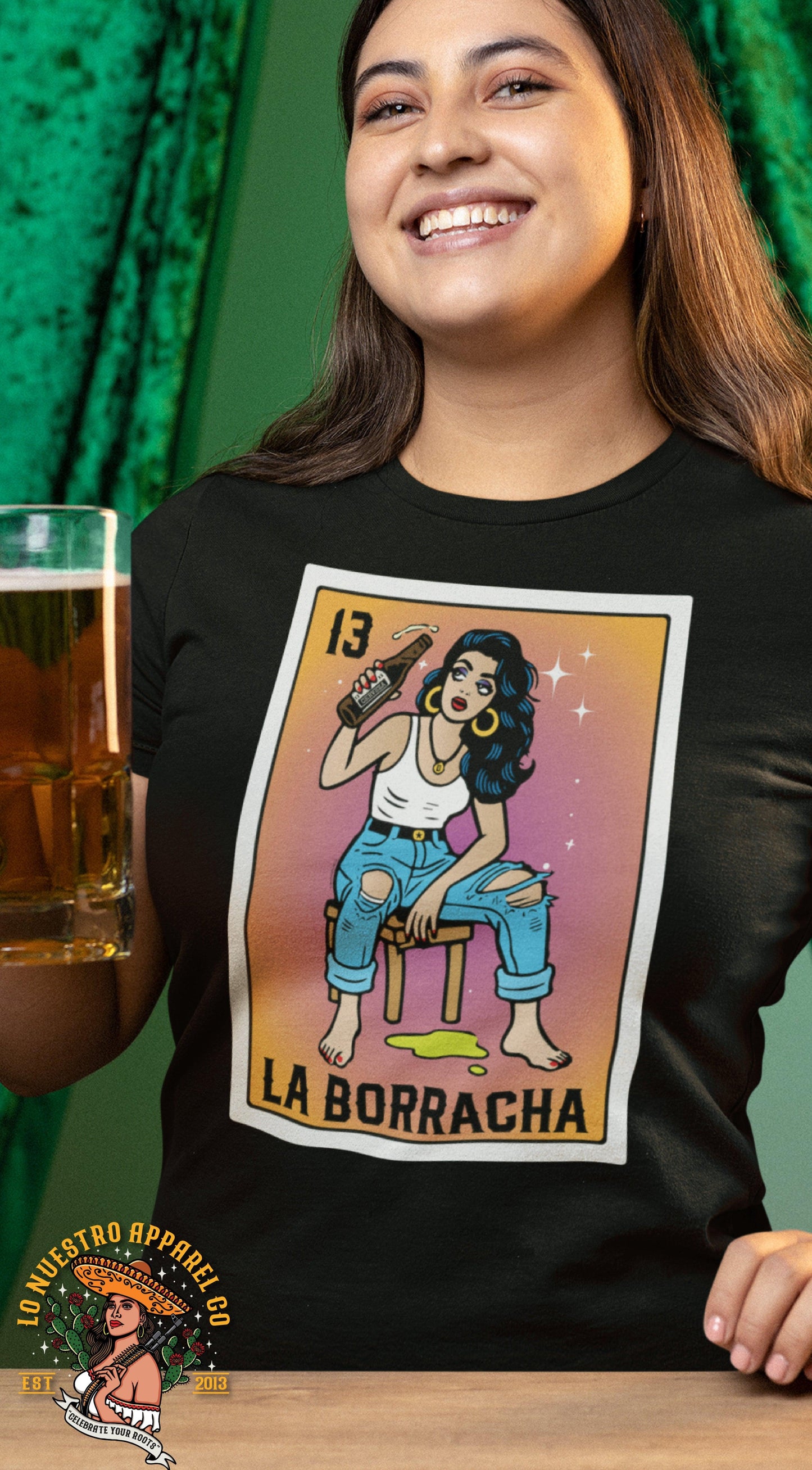 Model wearing the La Borracha Lotería T-Shirt while holding a beer mug. A Chicana-style tee inspired by the Mexican card game, perfect for fiestas and carne asadas.