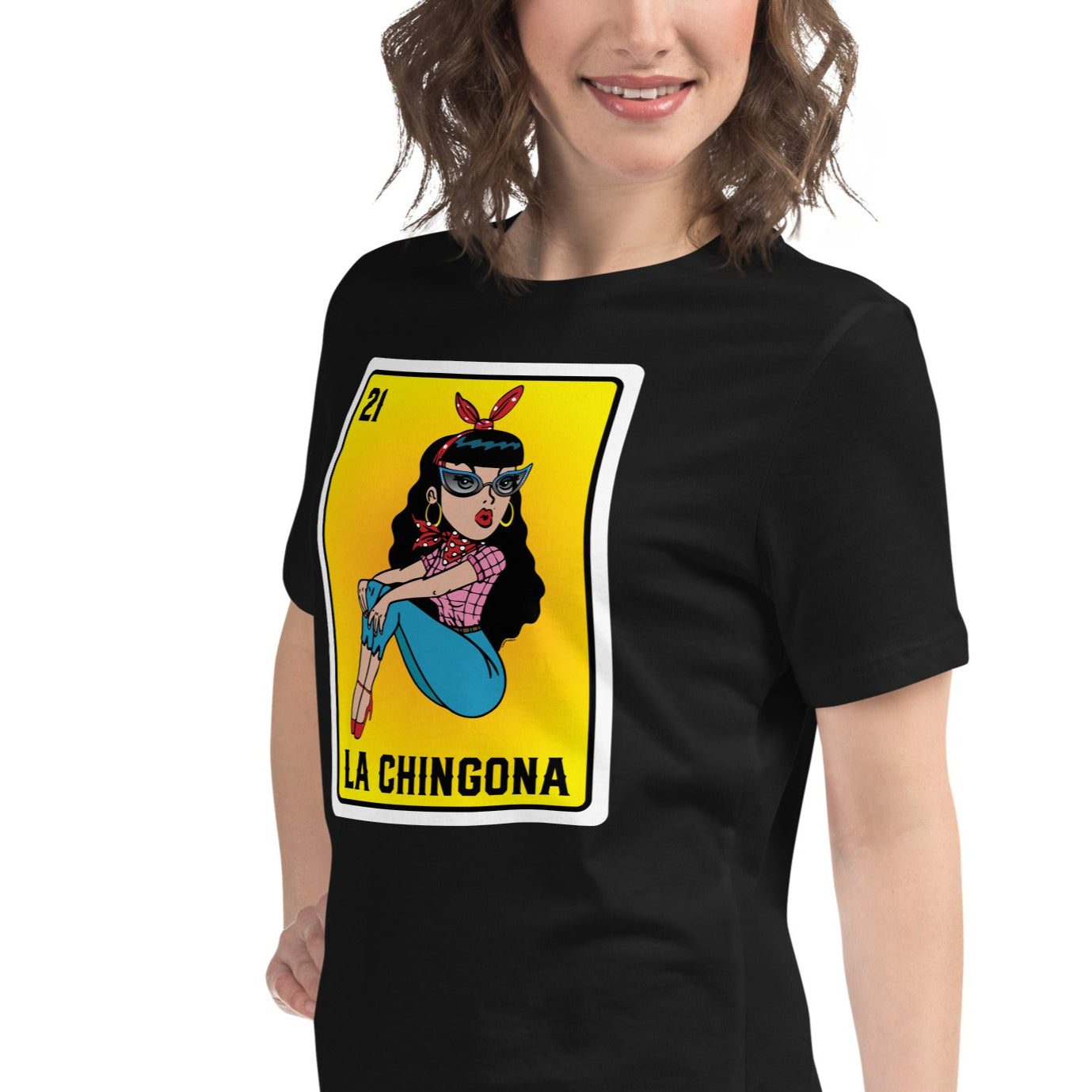 A young woman wearing a black t-shirt with a bold "La Chingona" Lotería-inspired design. The graphic features a Chicana in a pink plaid shirt, jeans, and high heels, sitting confidently against a vibrant orange-to-yellow gradient background.