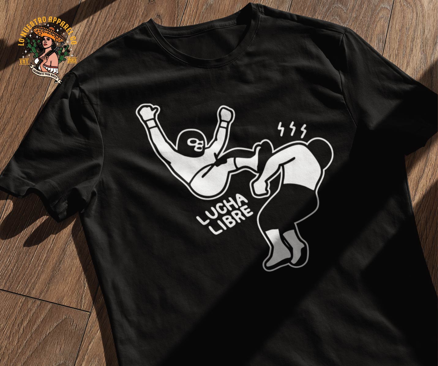 A wrinkled Lucha Libre Shirt Los Luchadores on a wooden floor with shadows and the Lo Nuestro Apparel Co logo.