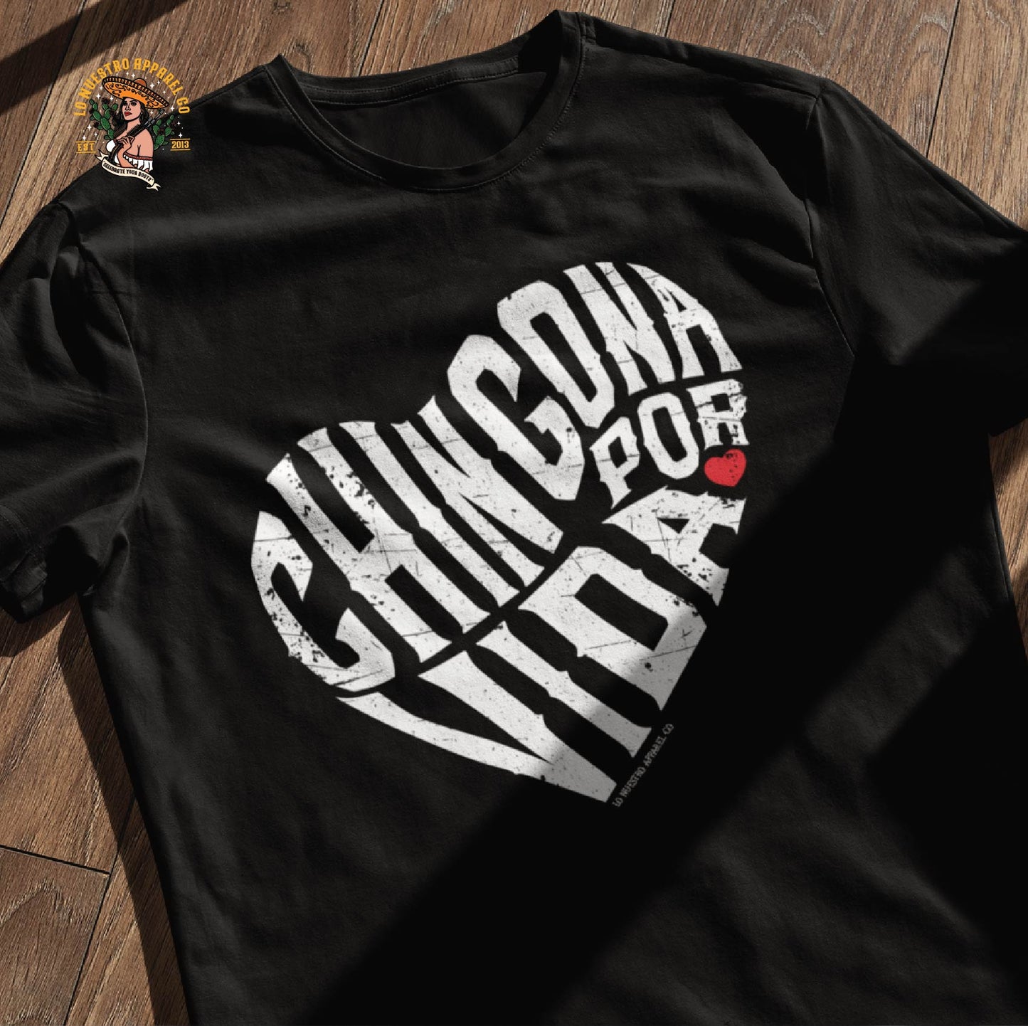 Black Chingona Por Vida Chicana shirt laid on a wooden surface, featuring a bold heart-shaped calligram design representing strength and cultura.