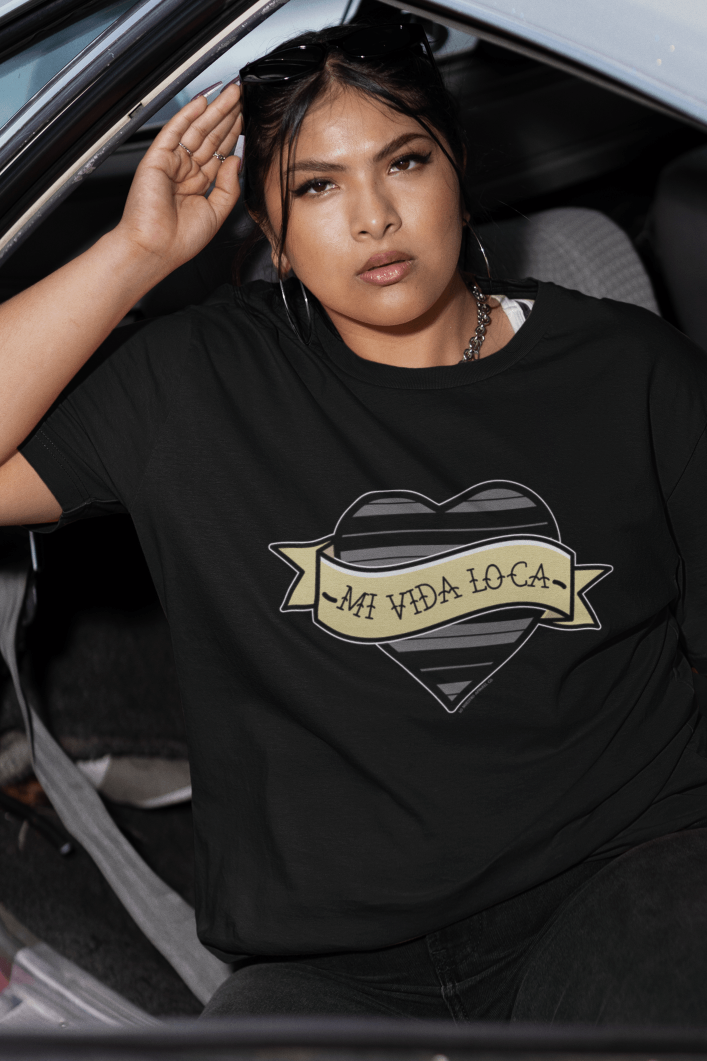 Chicana wearing the Mi Vida Loca Chola Heart T-Shirt inside a lowrider. Chola fashion with classic C Brown stripes. Perfect for cruising and car shows.