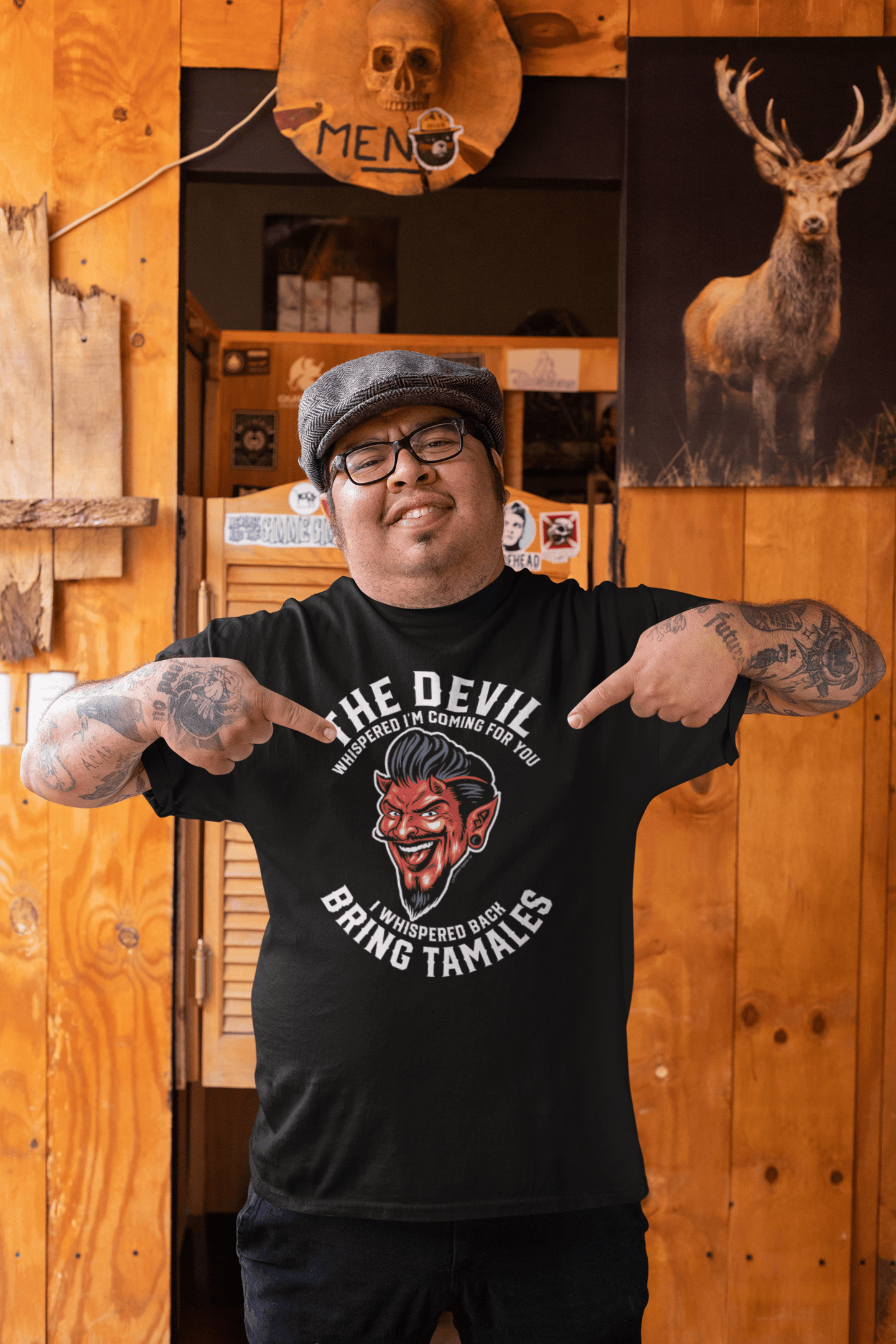 A man pointing at his black t-shirt in a rustic bar setting, featuring a devil graphic and the text 'The Devil Whispered I'm Coming For You. I Whispered Back Bring Tamales.
