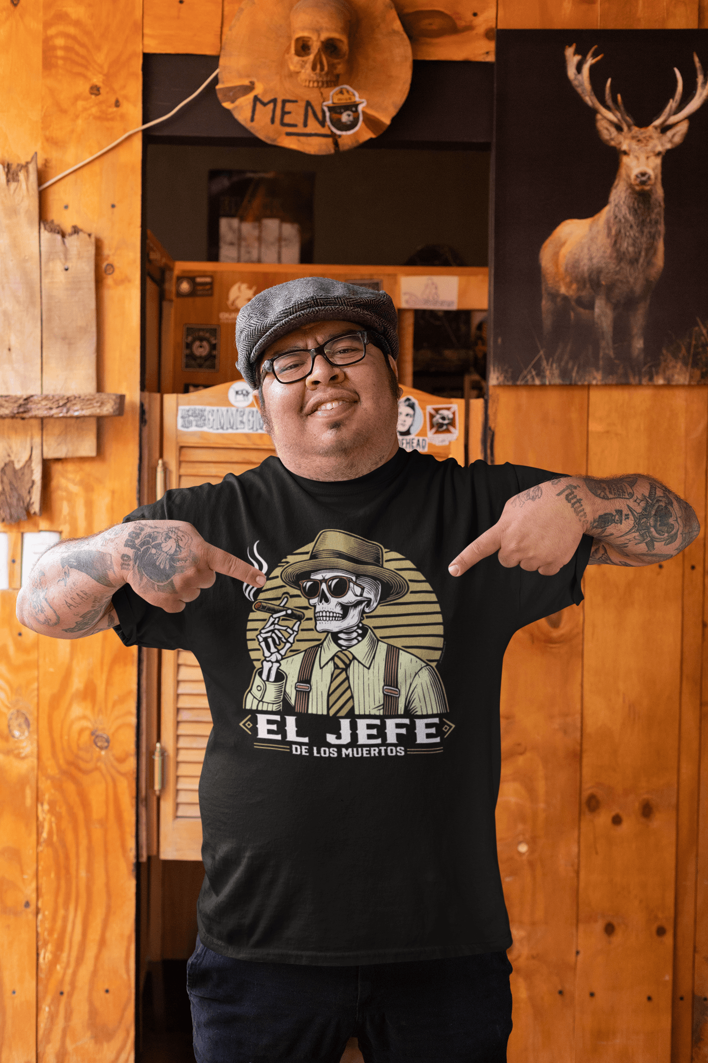 A man wearing the 'El Jefe de los Muertos' t-shirt while pointing to the design. The shirt features a skeleton in a fedora, sunglasses, and tie, holding a cigar. The background shows a rustic bar setting.