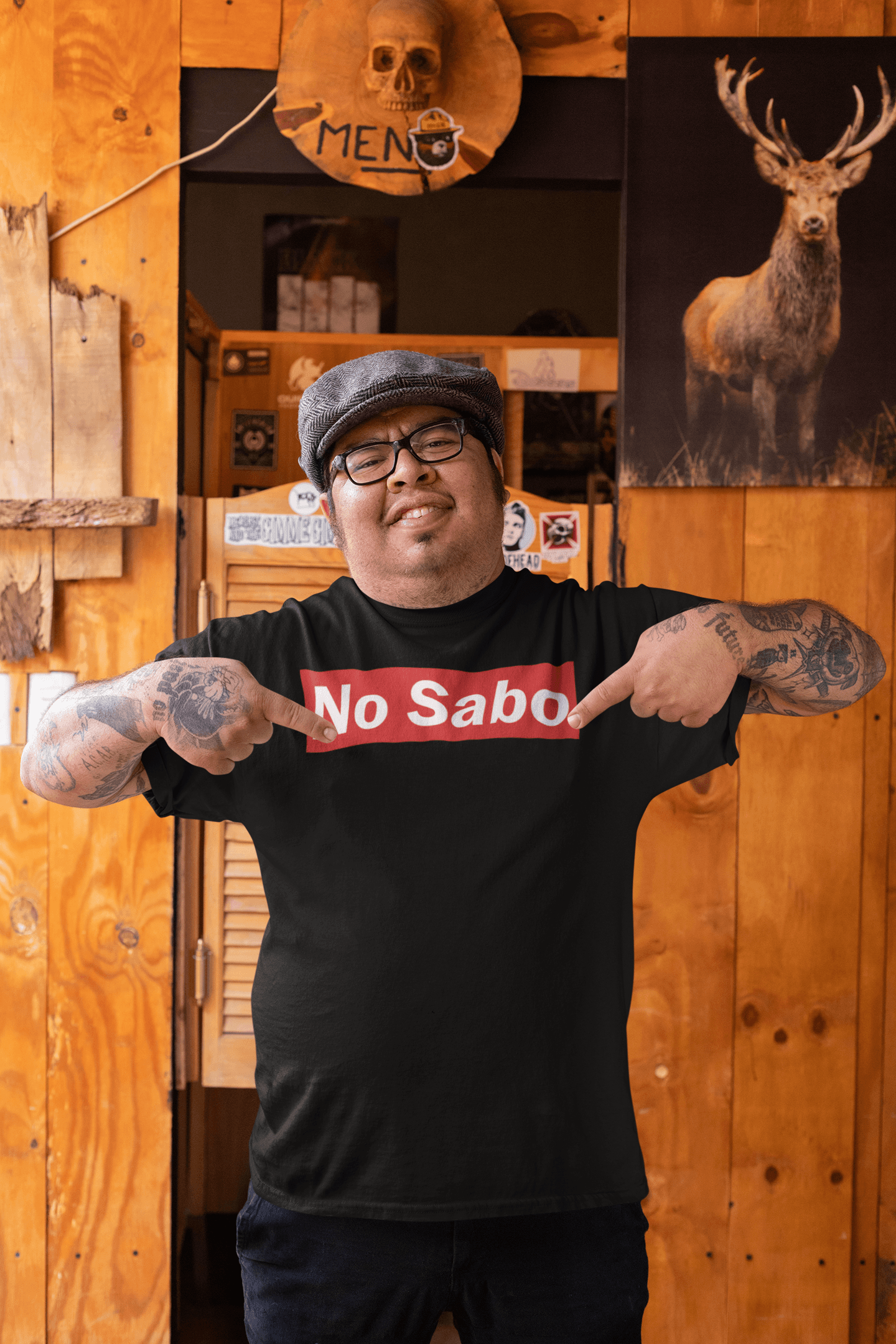 A man standing in a rustic bar setting, wearing a black t-shirt with the 'No Sabo' design in white text on a red rectangular background, pointing proudly at the design.