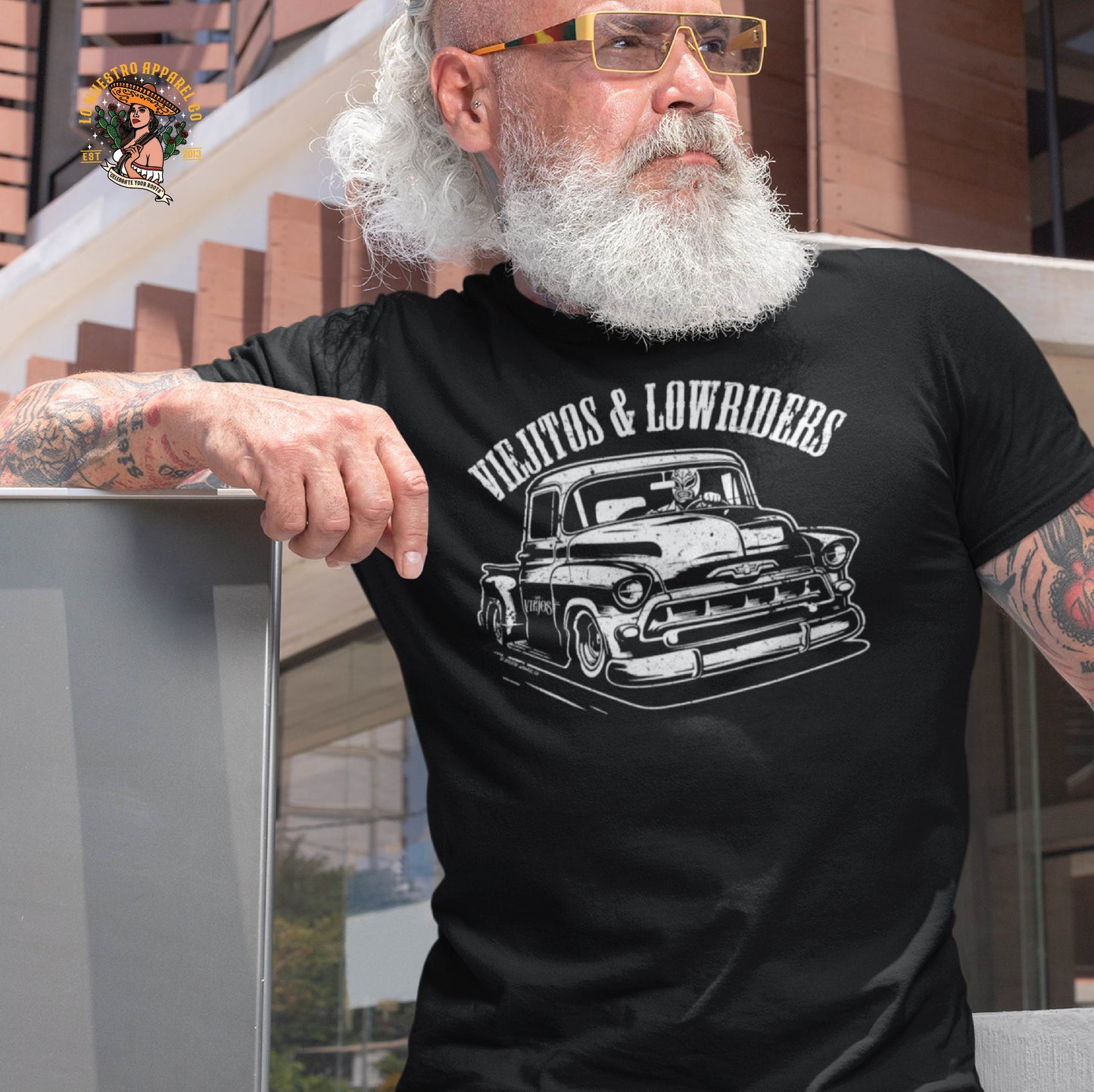 A stylish bearded man wearing the Viejitos & Lowriders lowrider t-shirt, featuring a 1957 Chevy Stepside and a luchador driver. This Chicano clothing piece is perfect for car shows, cruising, and those who live the lowrider lifestyle.