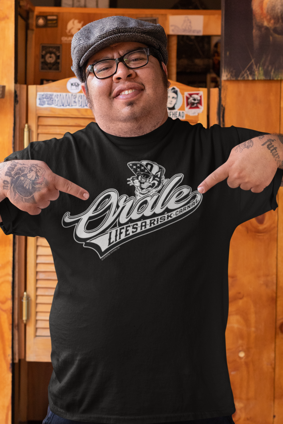 Man wearing a black "Órale Life's a Risk Carnal" T-shirt, pointing at the bold baseball-style graphic featuring an angry cat in a bandana.