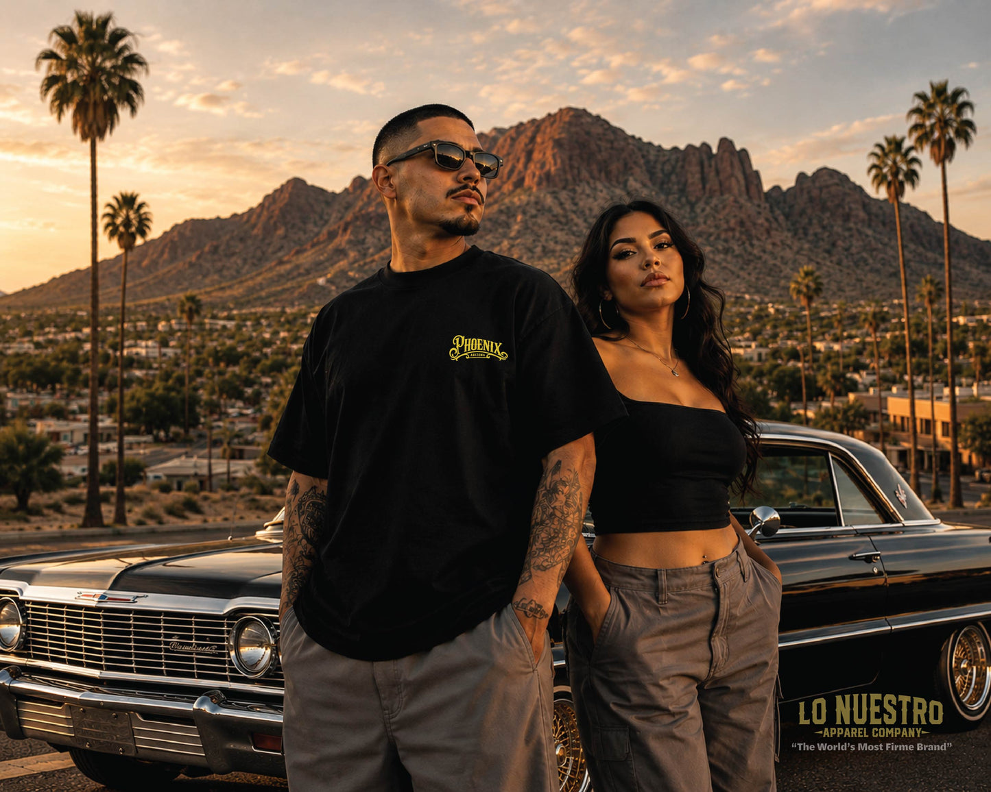Back view of the Phoenix California plaque T-shirt worn by a Hispanic man beside a woman with Camelback Mountain and a classic black 1963 Chevrolet Impala in the background.