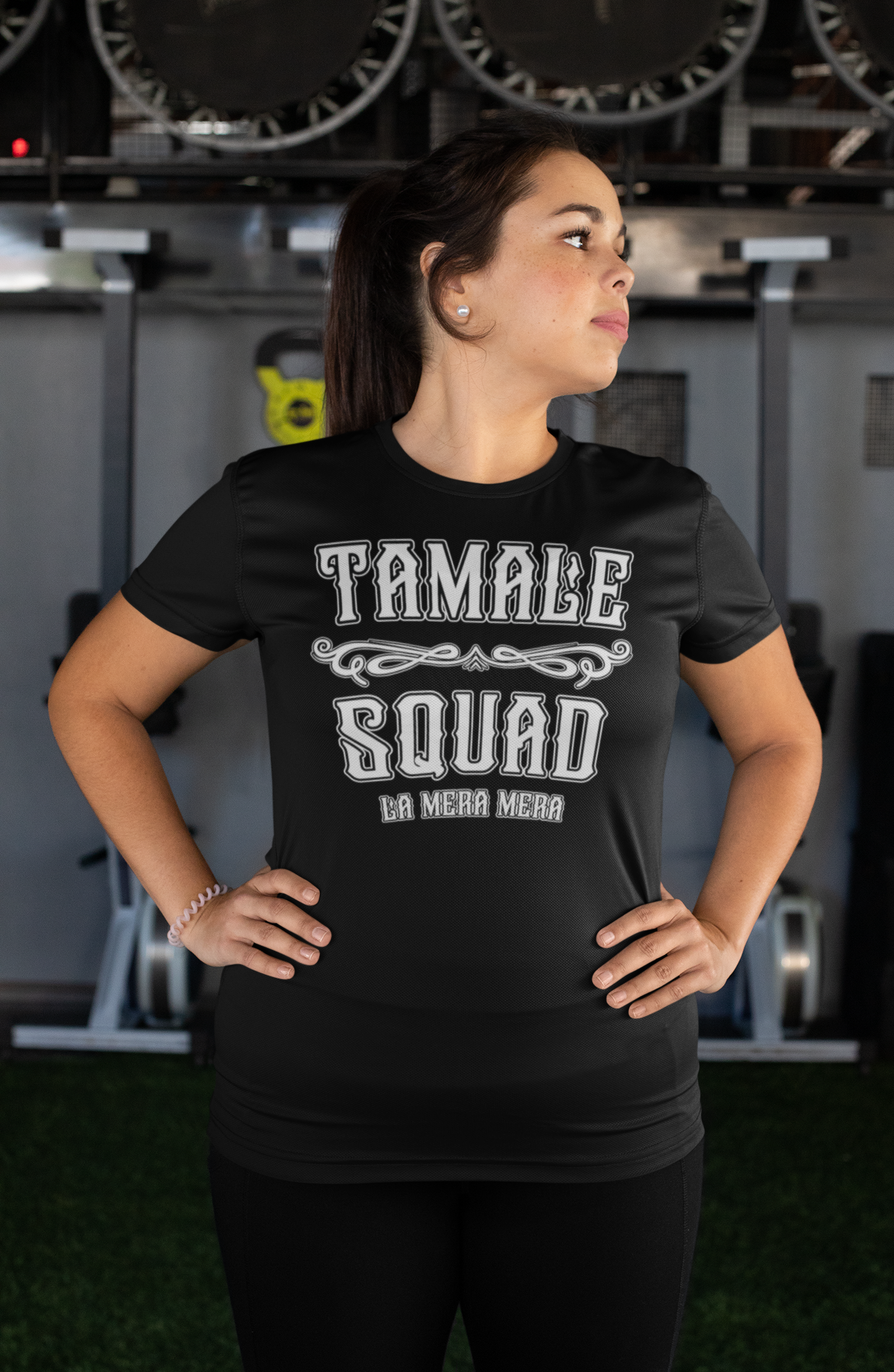 Woman wearing a black "Tamale Squad Women's T-Shirt" with a front graphic that reads "Tamale Squad" and "La Mera Mera," standing confidently.