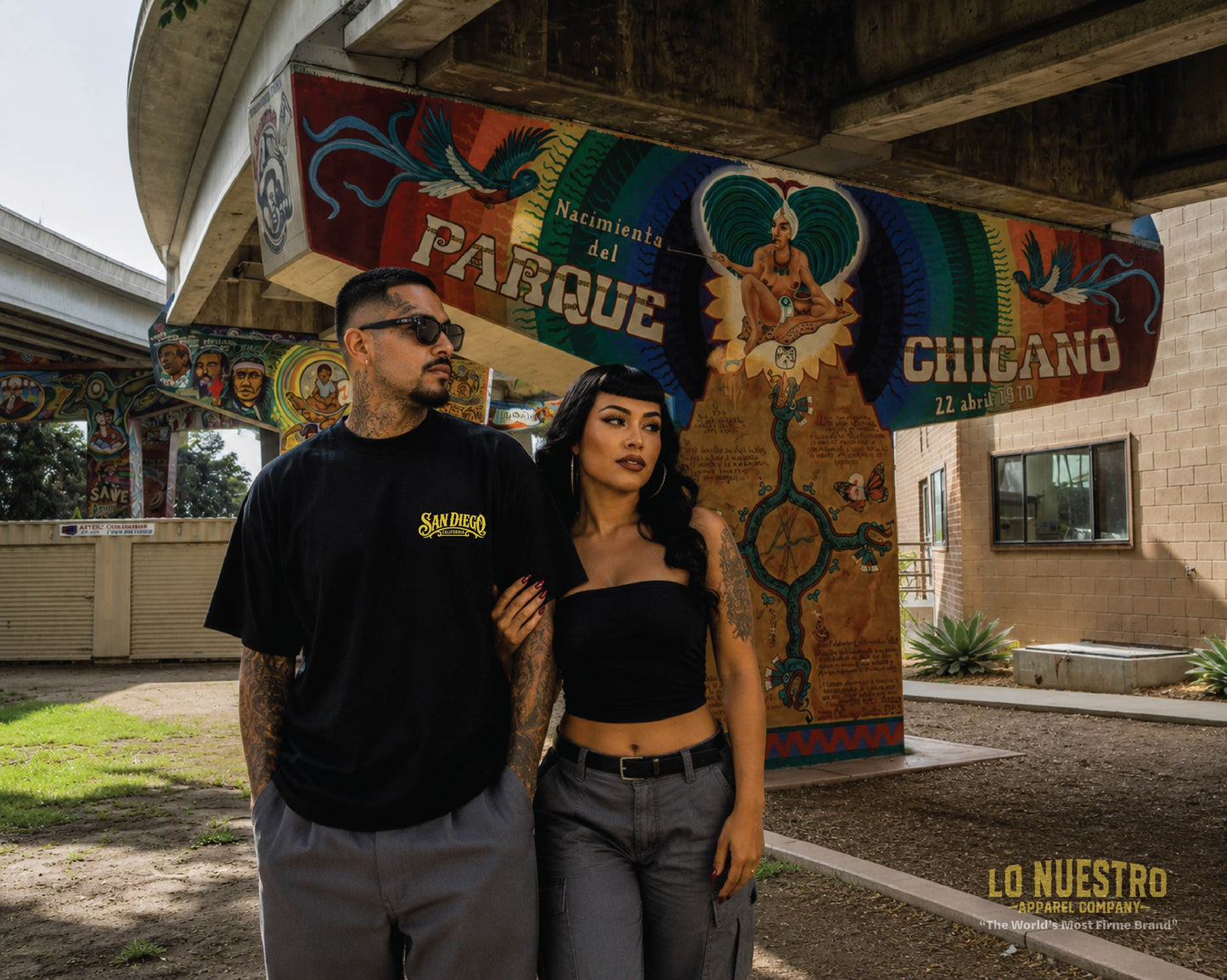 Man and woman wearing the San Diego Lowrider Plaque T-shirt in Chicano Park with the iconic murals beneath the Coronado Bridge in San Diego.