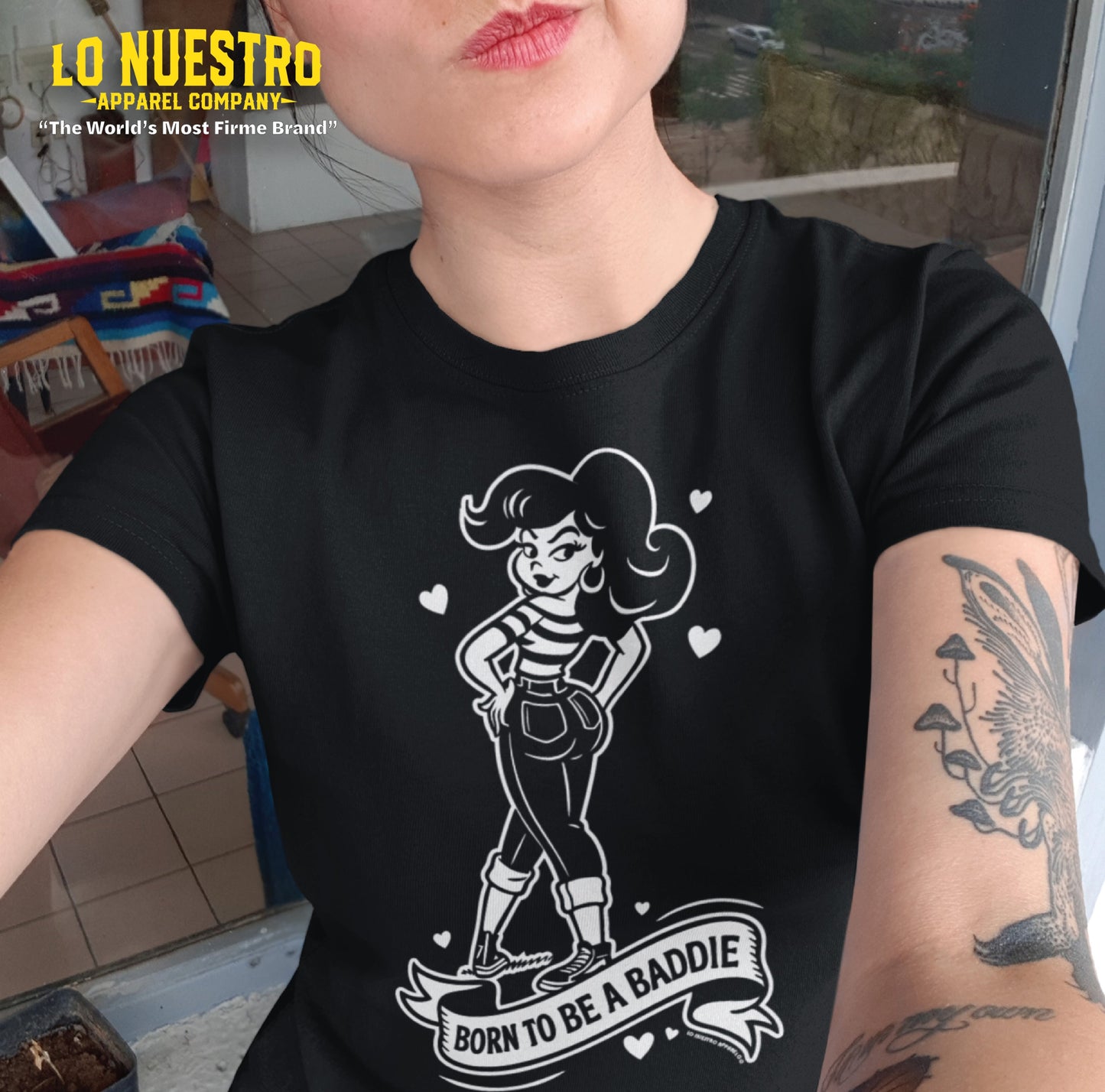 Tattooed woman taking a selfie wearing a black Chicana shirt that says Born to Be a Baddie with a cartoon graphic.