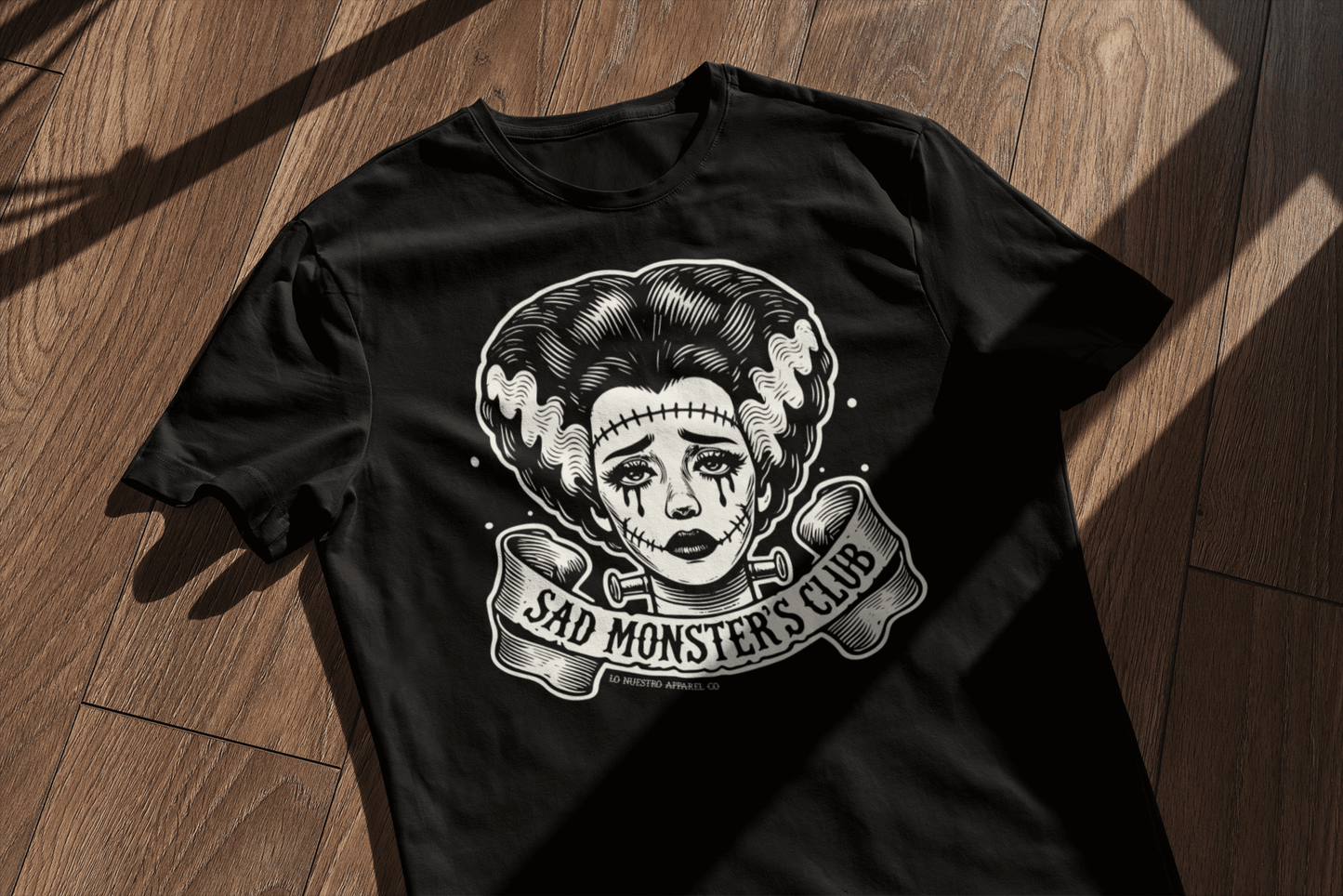 Black t-shirt with a retro tattoo-style 'Sad Monster's Club' Bride of Frankenstein design, laid flat on a wooden surface with window shadows adding a dramatic effect.