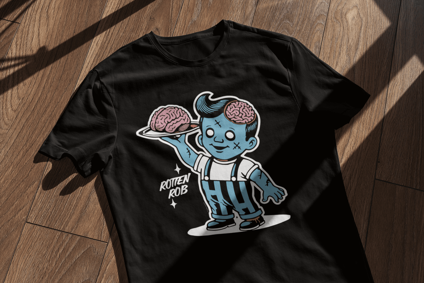 A black t-shirt featuring the 'Rotten Rob' zombie design, laid flat on a wooden surface with window shadows. The retro Halloween style stands out with the character holding a brain on a platter.