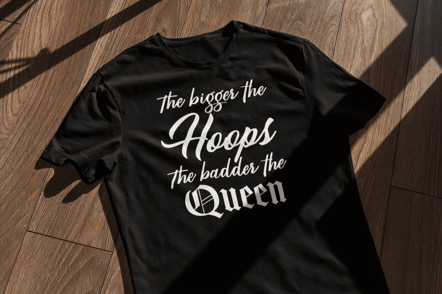 A black t-shirt laid flat on a wooden surface with sunlight casting a window-shaped shadow. The shirt reads 'The bigger the Hoops, the badder the Queen' in a combination of cursive and gothic fonts, giving off a bold and empowering vibe