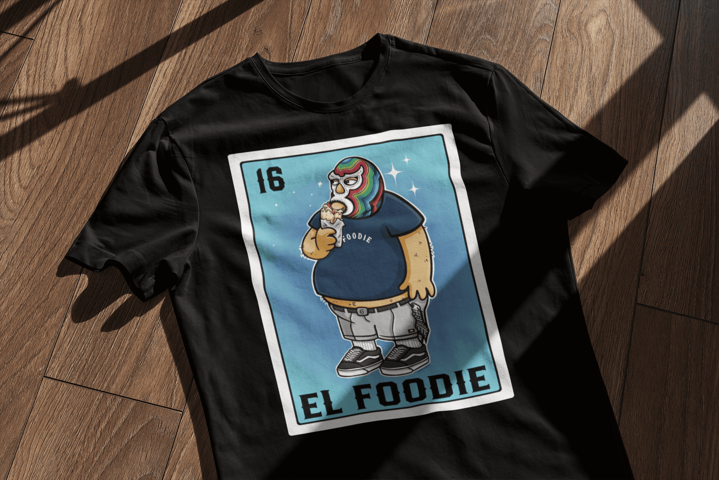 Flat lay of the 'El Foodie' T-shirt on a wooden floor, showcasing a Lotería-style design of a luchador holding a burrito.