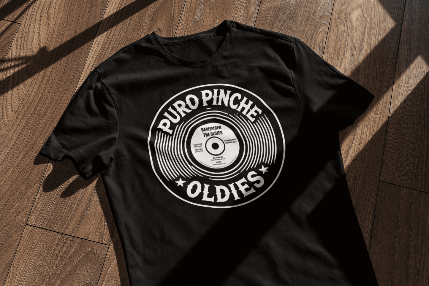 A black graphic tshirt with the text Puro Pinche Oldies surrounded by a vinyl record laying on a wood floor.