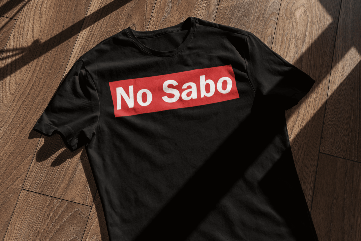 Black t-shirt featuring the bold 'No Sabo' design in white text on a red rectangular background, displayed on a wooden surface with sunlight casting shadows