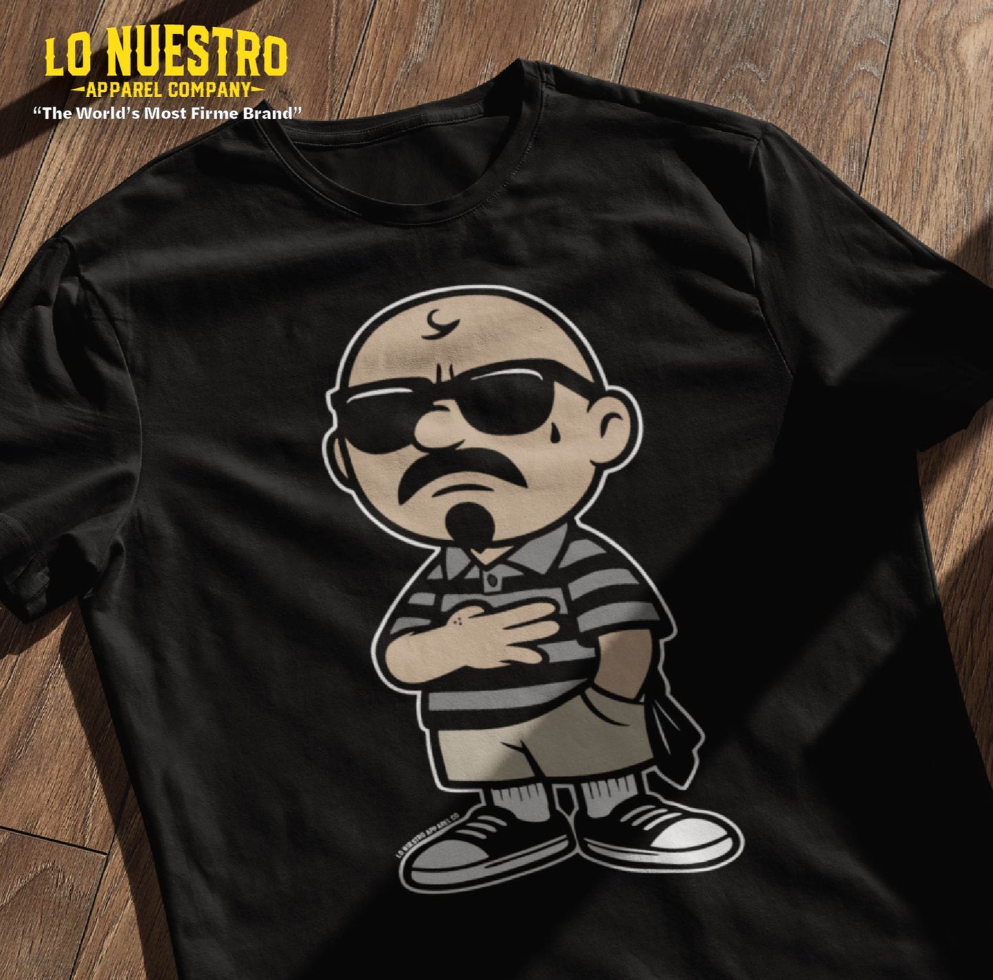 Black t-shirt mockup on a wooden floor featuring a cartoon homie design. A funny Mexican shirt styled with Chicano clothing influence.