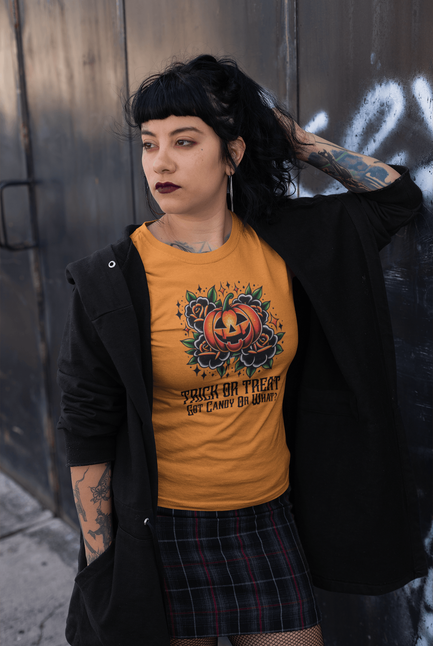 Goth woman with tattoos leaning against a wall, wearing a Heather Marmalade 'Trick or Treat, Got Candy Or What?' t-shirt.