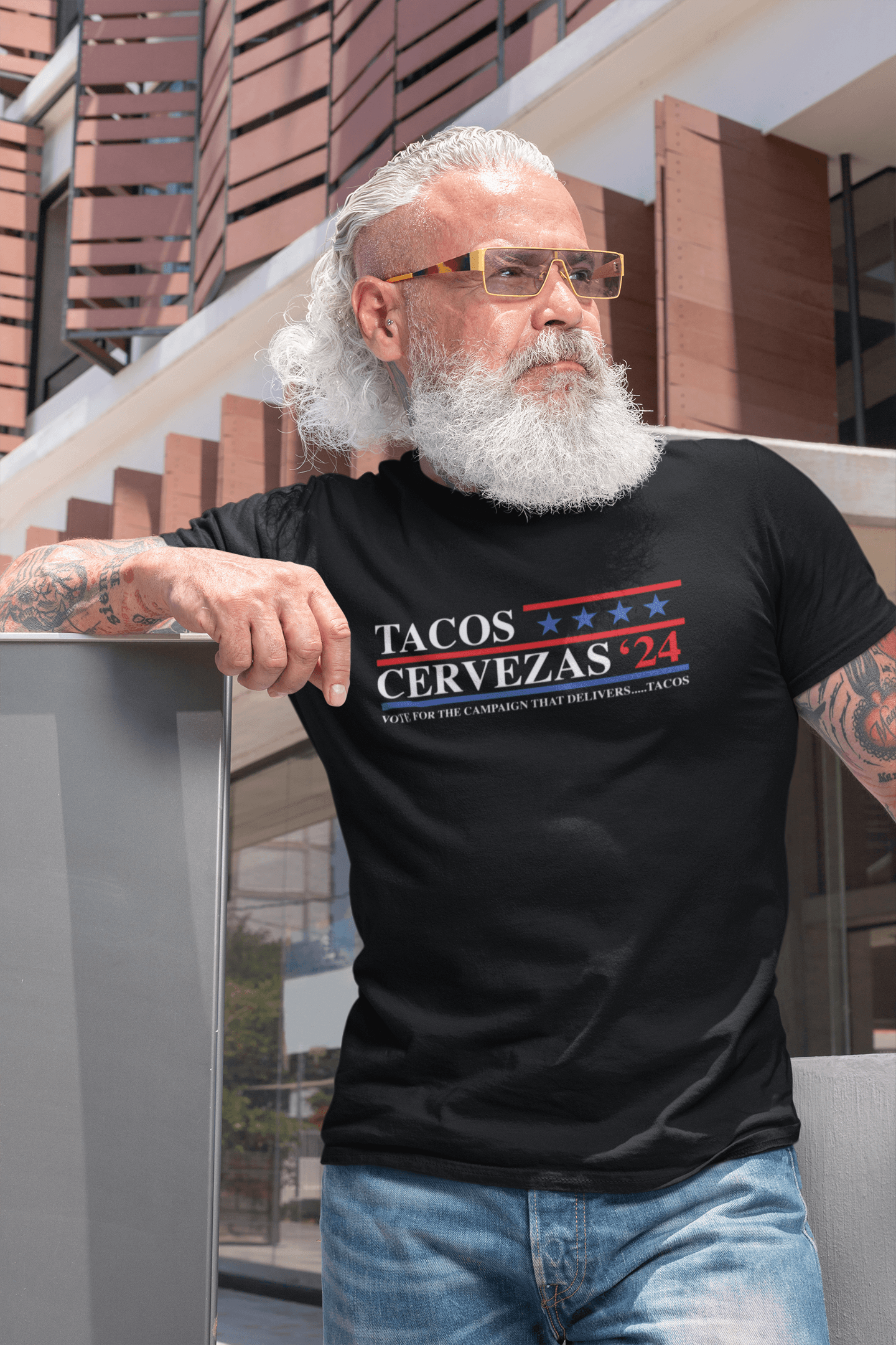 Middle-aged man with a white beard and sunglasses wearing a black T-shirt with the slogan 'Tacos & Cervezas '24 - Vote for the campaign that delivers... tacos' standing outdoors.