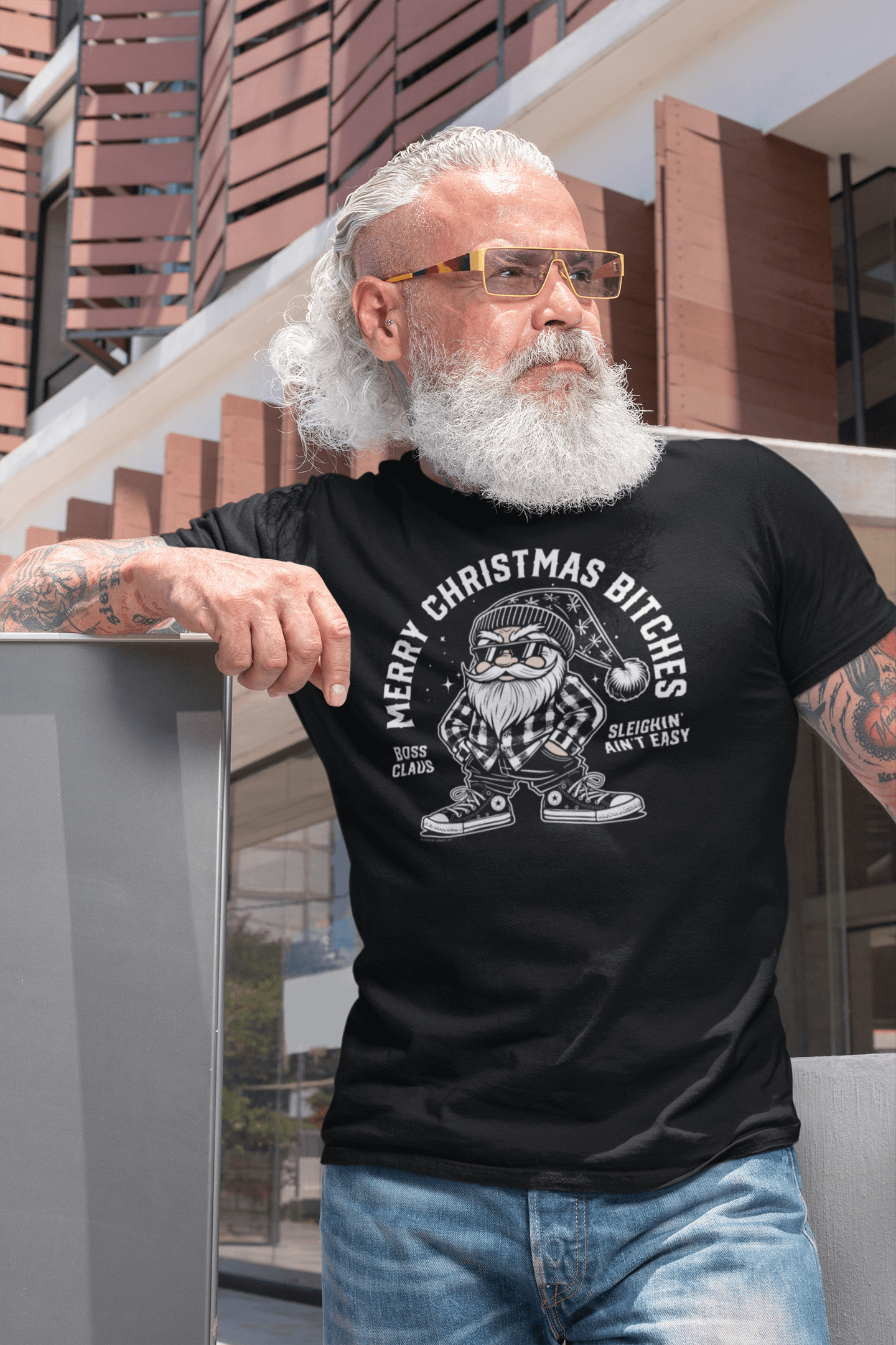 Trendy middle-aged man with white hair and sunglasses wearing a black t-shirt that reads 'Merry Christmas Bitches,' showcasing a street-style Santa in a plaid shirt with a cholo look, standing outside a modern building.