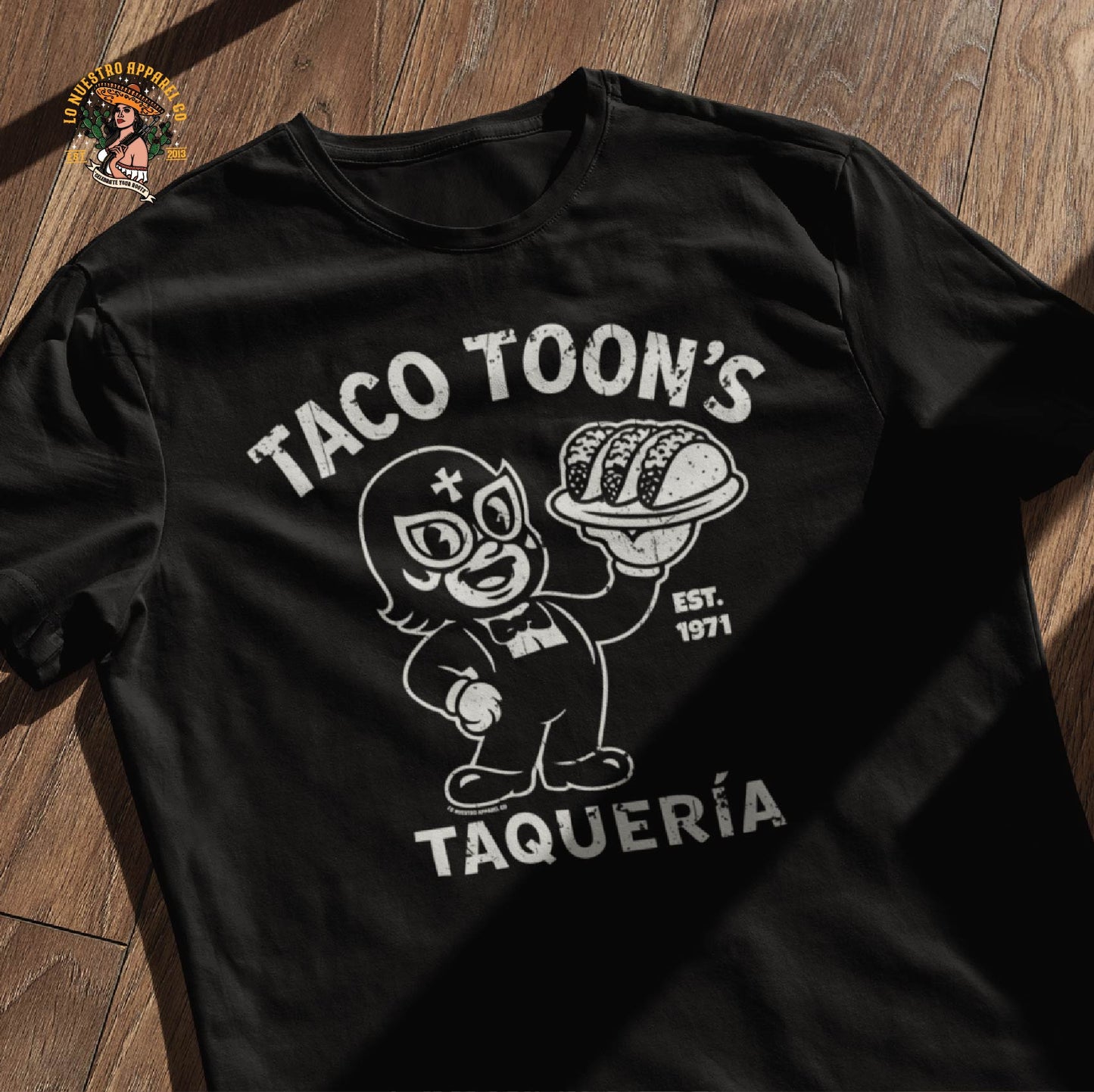 Taco Toon’s Taquería funny Mexican shirt laid flat on a wooden background. Vintage cartoon luchador design perfect for fans of tacos and funny shirts.