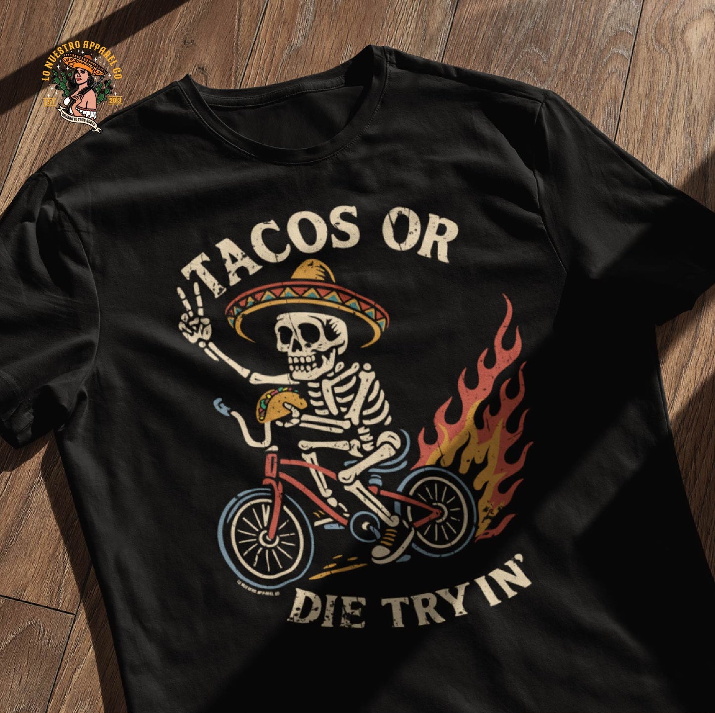 Tacos Or Die Tryin design on a black t-shirt, laid flat on a wooden floor. A bold graphic perfect for fans of Chicano shirts and funny shirts with attitude.