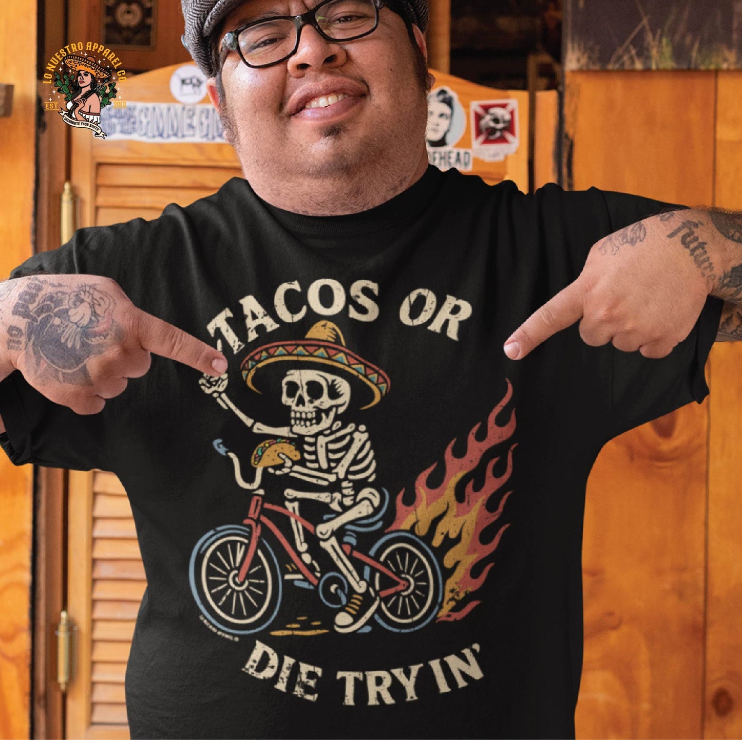 Man wearing the Tacos Or Die Tryin Chicano shirt inside a rustic taquería. This funny shirt features a skeleton on a lowrider bike, bringing cultura and humor together.