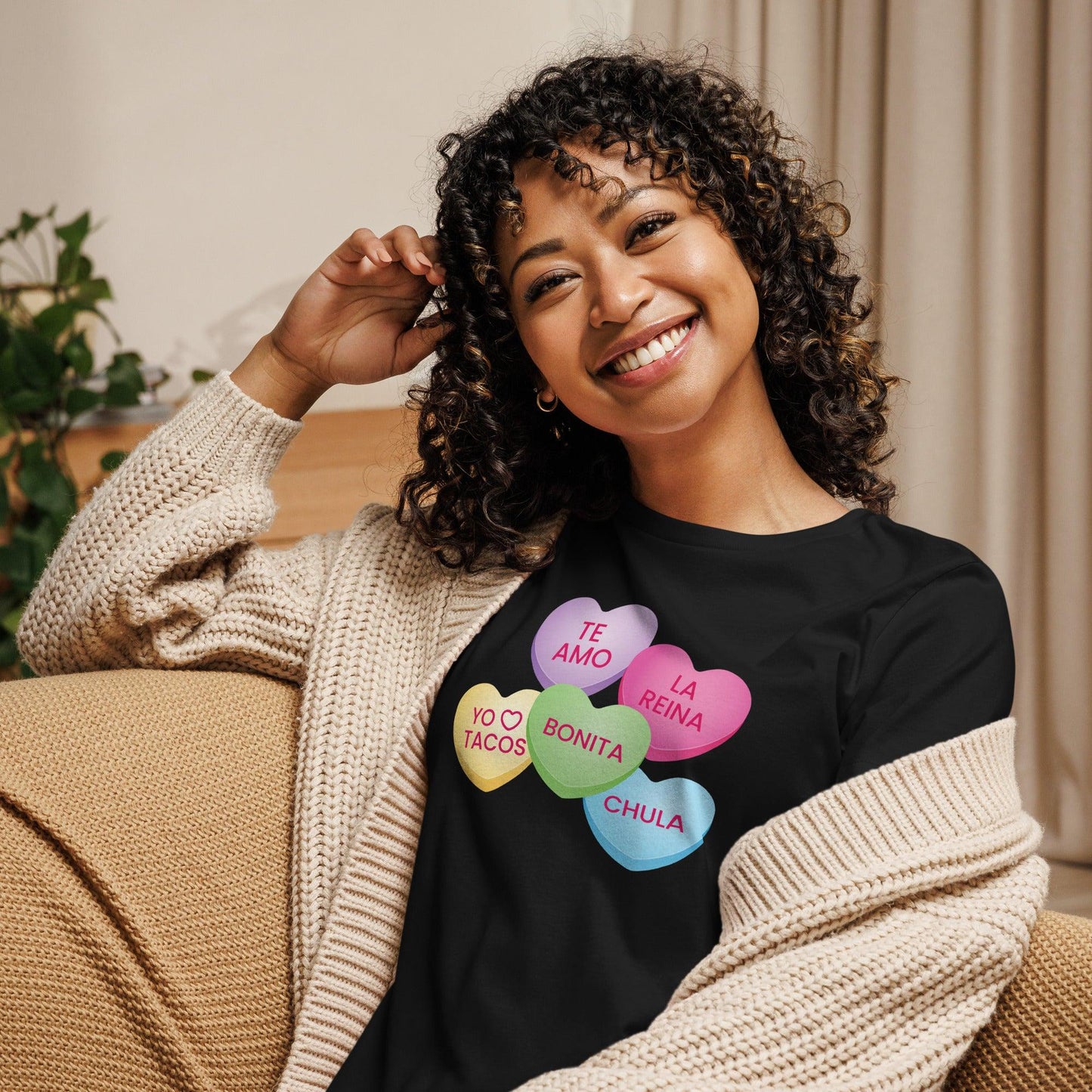 A young woman sitting on a couch wearing a knitted sweater with a black tshirt with traditional Valentin's day hearts with Spanish text.